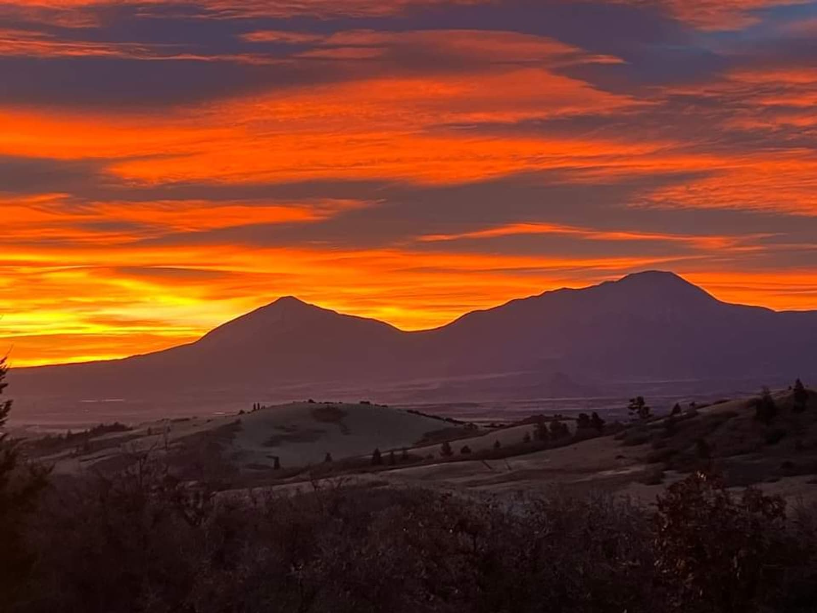 Vivid orange and yellow sunset sky over two mountain peaks, with rolling hills and trees in the foreground.