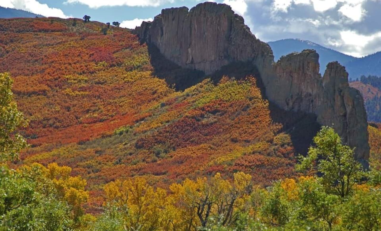 A rocky ridge cuts through a hillside covered in vibrant autumn foliage, with red, yellow, and orange leaves. Trees frame the foreground, and cloudy mountains rise in the background.