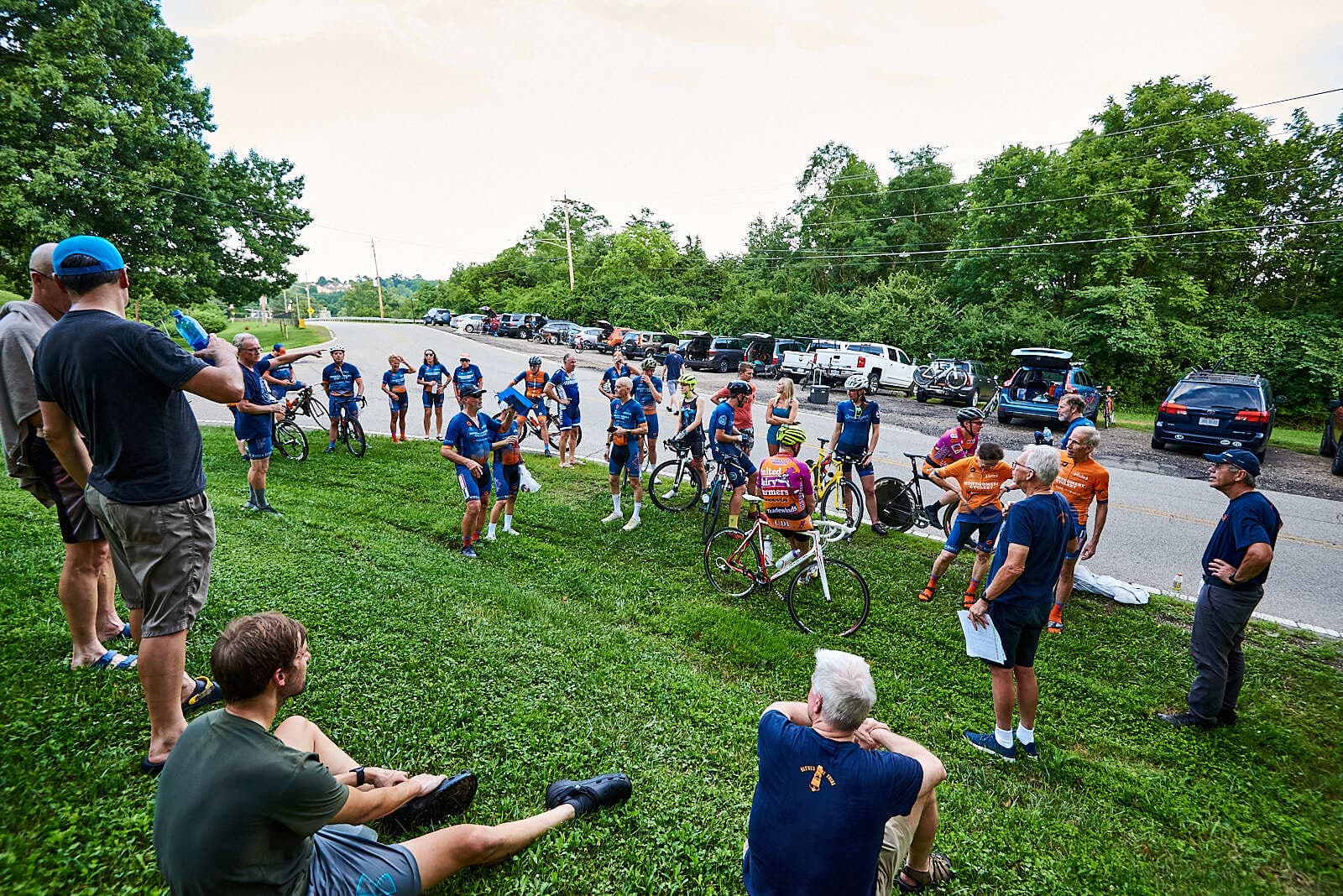 A group of cyclists in colorful jerseys gathers on a grassy area near parked cars as people sit and stand, listening to someone speaking. Trees and a road are visible in the background.