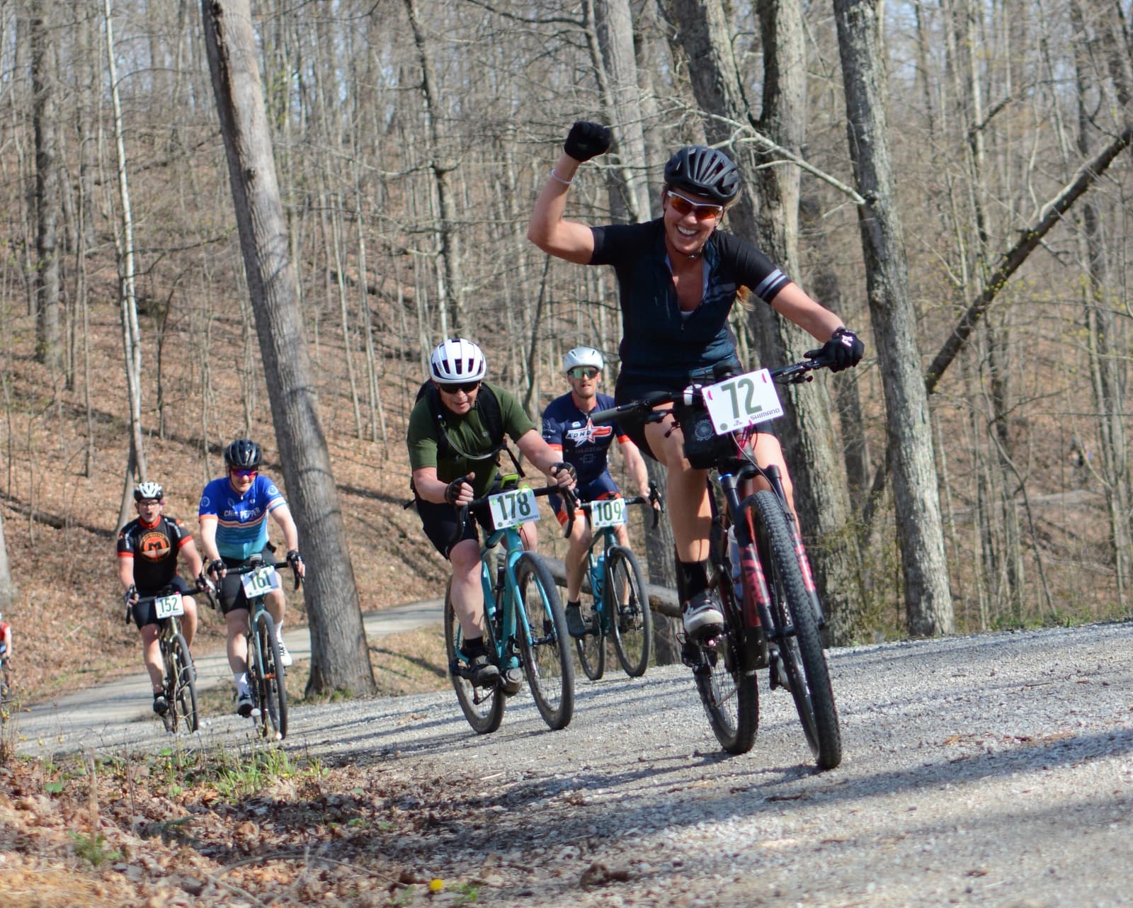 A group of cyclists race uphill on a gravel road in a forest. The leading cyclist, wearing black and number 72, raises a fist triumphantly while others pedal behind, surrounded by leafless trees.