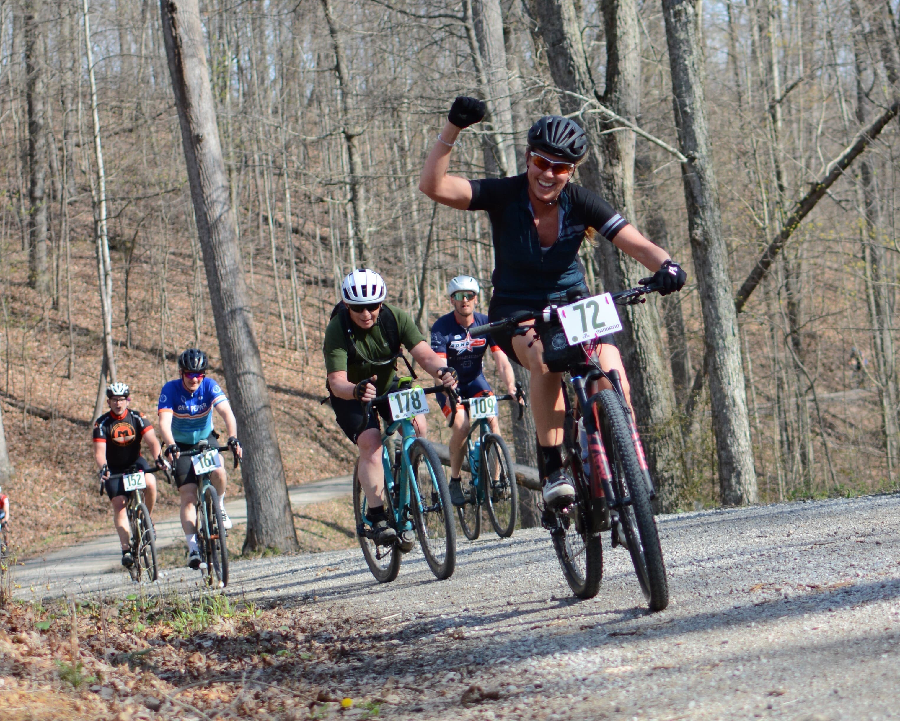 A group of cyclists race uphill on a gravel road in a forest. The leading cyclist, wearing black and number 72, raises a fist triumphantly while others pedal behind, surrounded by leafless trees.