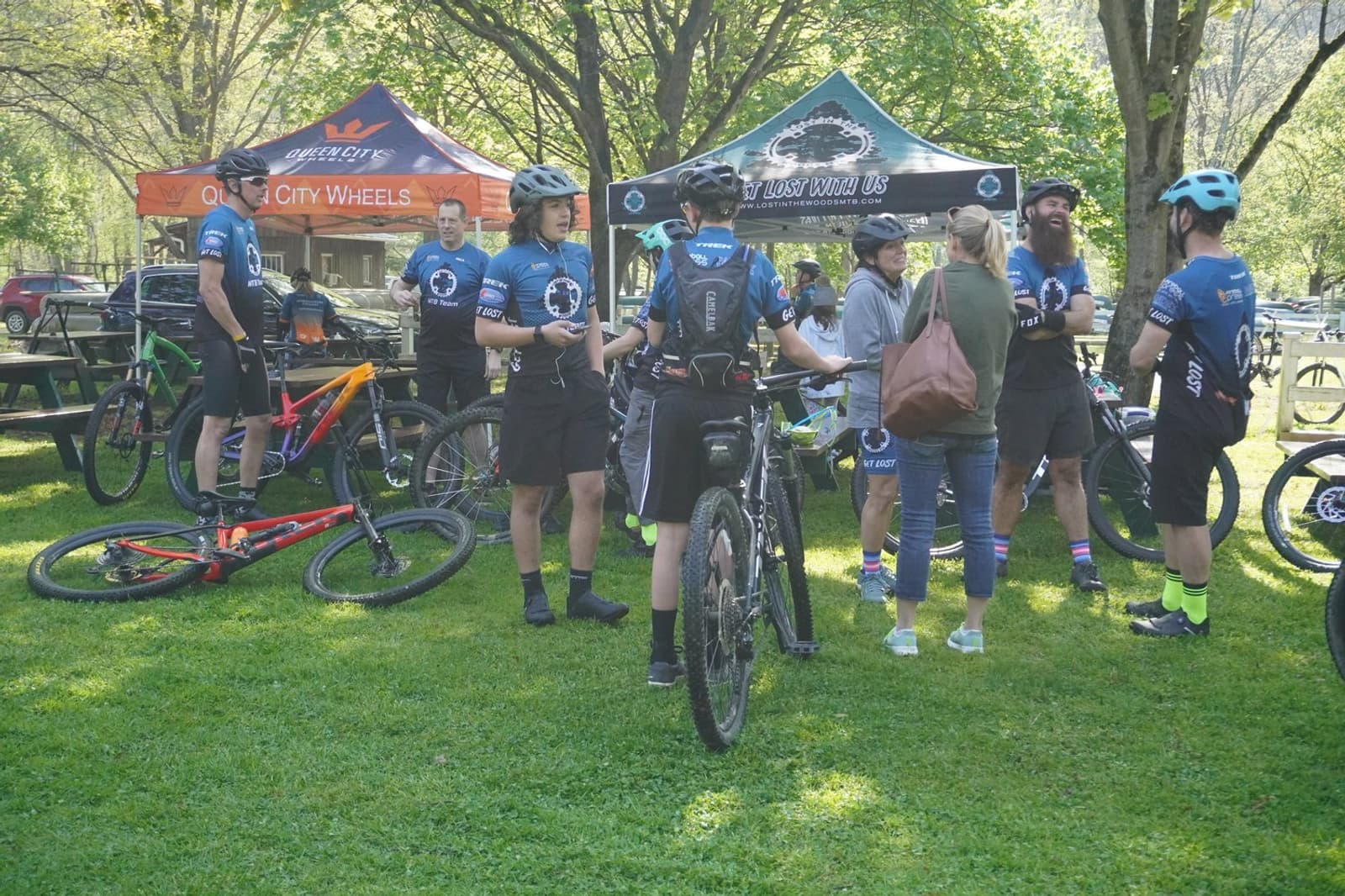 A group of cyclists in matching blue jerseys stand and chat near tents, parked bikes, and picnic tables on a grassy lawn, with trees and sunlight in the background.