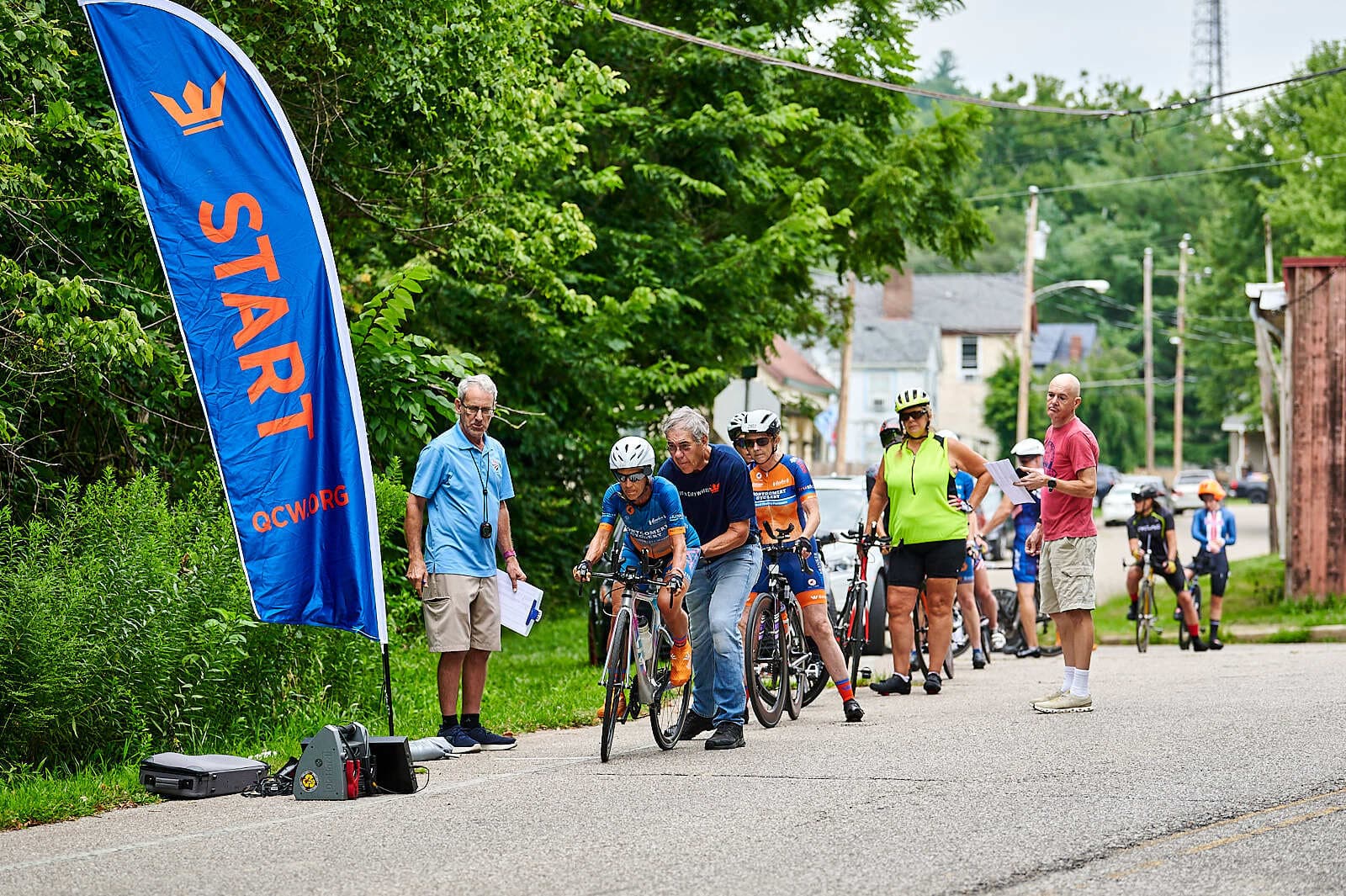 Cyclists line up at a race start, with volunteers assisting. A blue "START" flag stands beside them. Trees and buildings are visible in the background on a cloudy day.