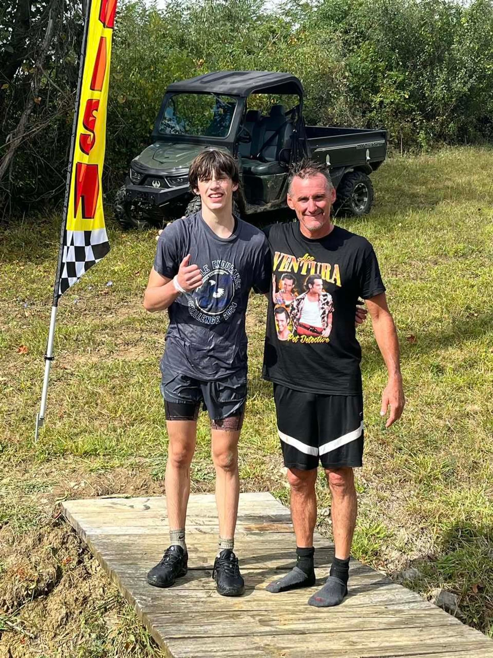 Two people, one younger and one older, stand on a wooden path outdoors, smiling at the camera. The younger person is wet and giving a thumbs up. A flag and an off-road vehicle are in the background, with trees behind them.