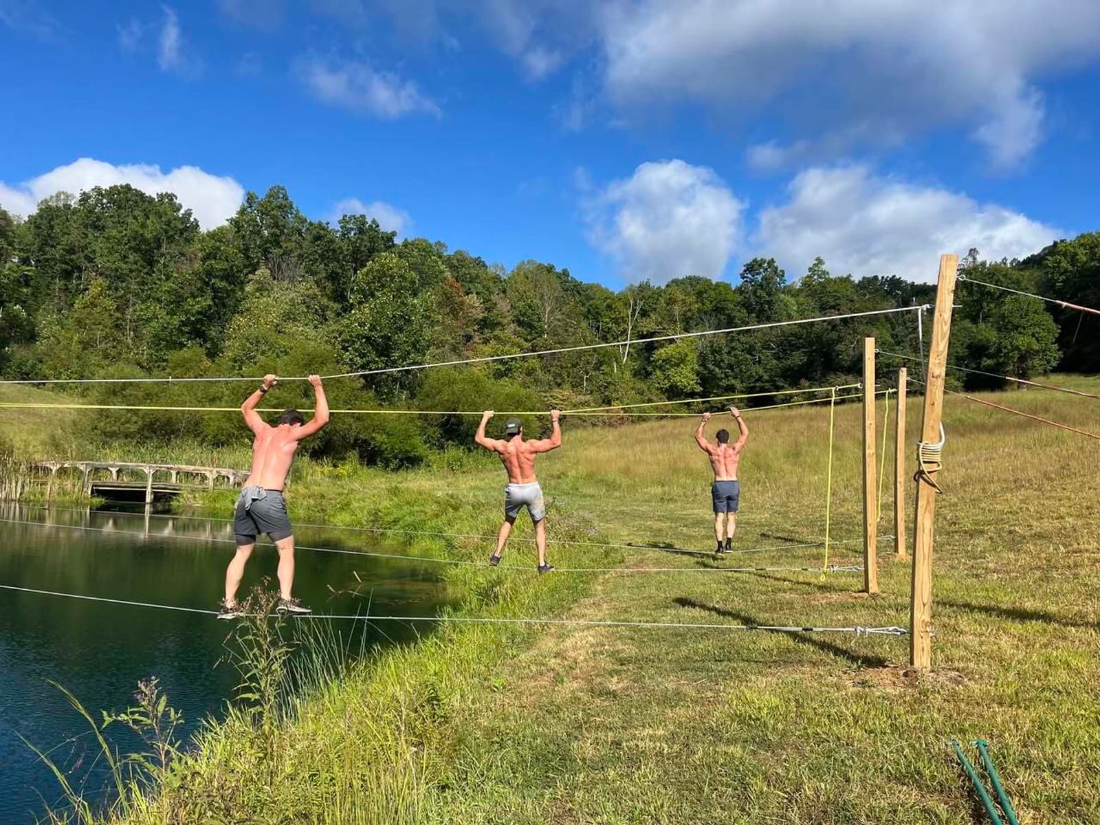Three shirtless men in shorts cross a pond using ropes suspended between wooden posts, with grassy hills and trees in the background under a blue sky with clouds.