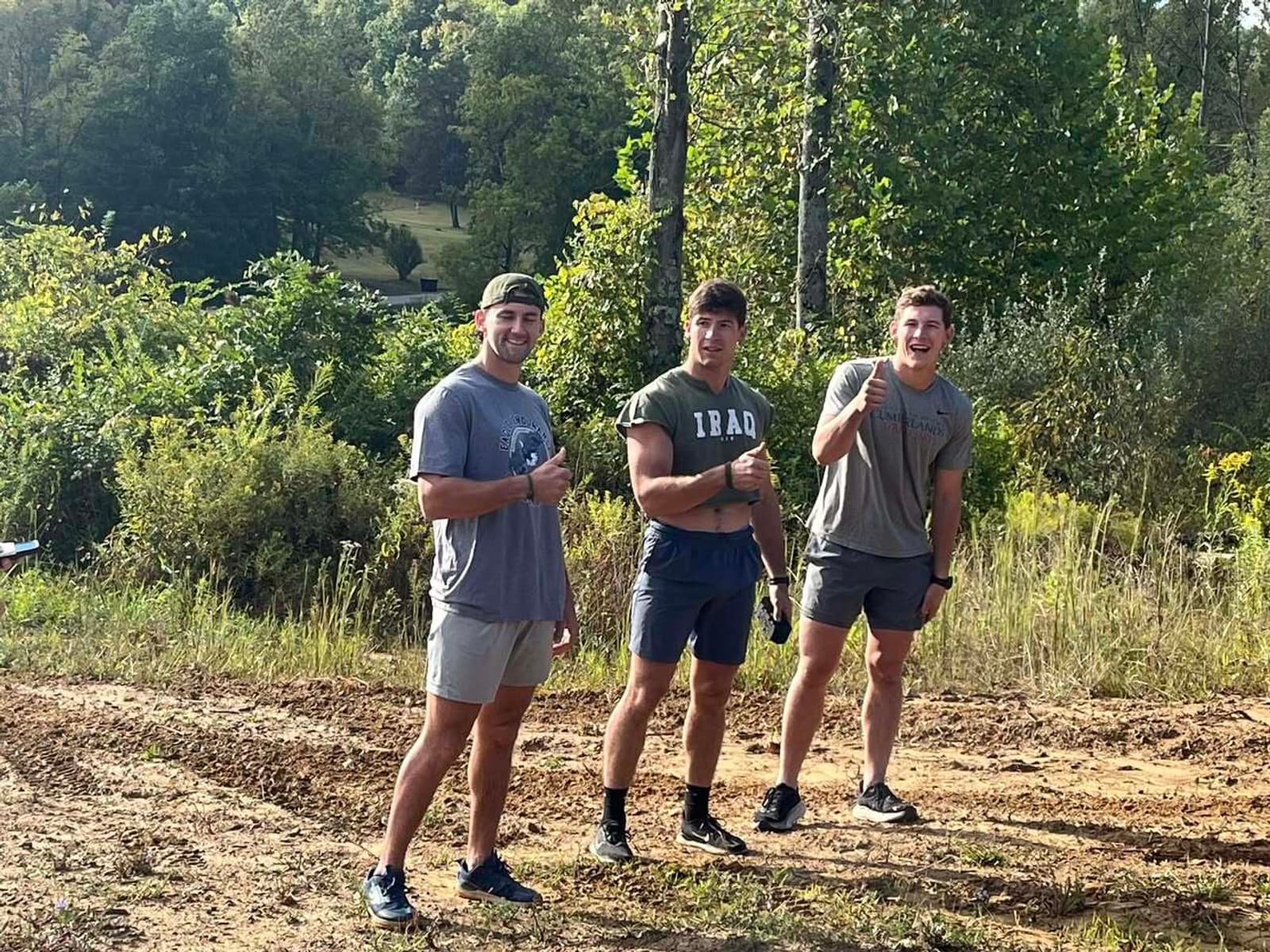Three young men stand outdoors on a dirt path, smiling and giving thumbs up. They are dressed in athletic wear and surrounded by green trees and vegetation on a sunny day.