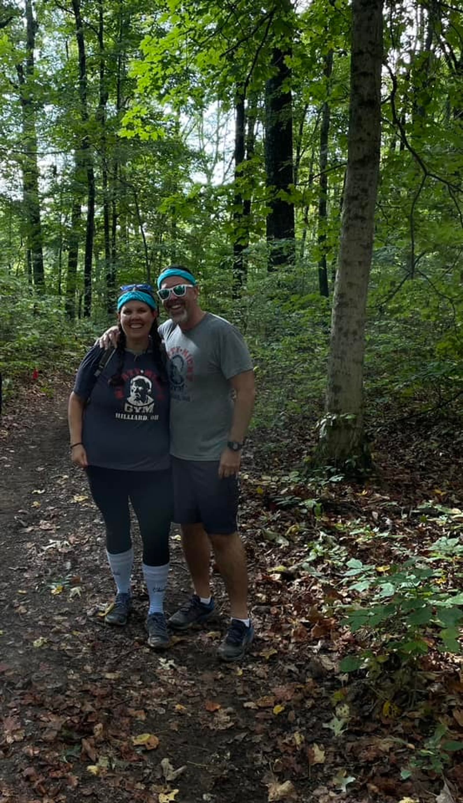 Two people wearing athletic clothes and headbands stand smiling together on a wooded trail, surrounded by lush green trees and fallen leaves on the ground.