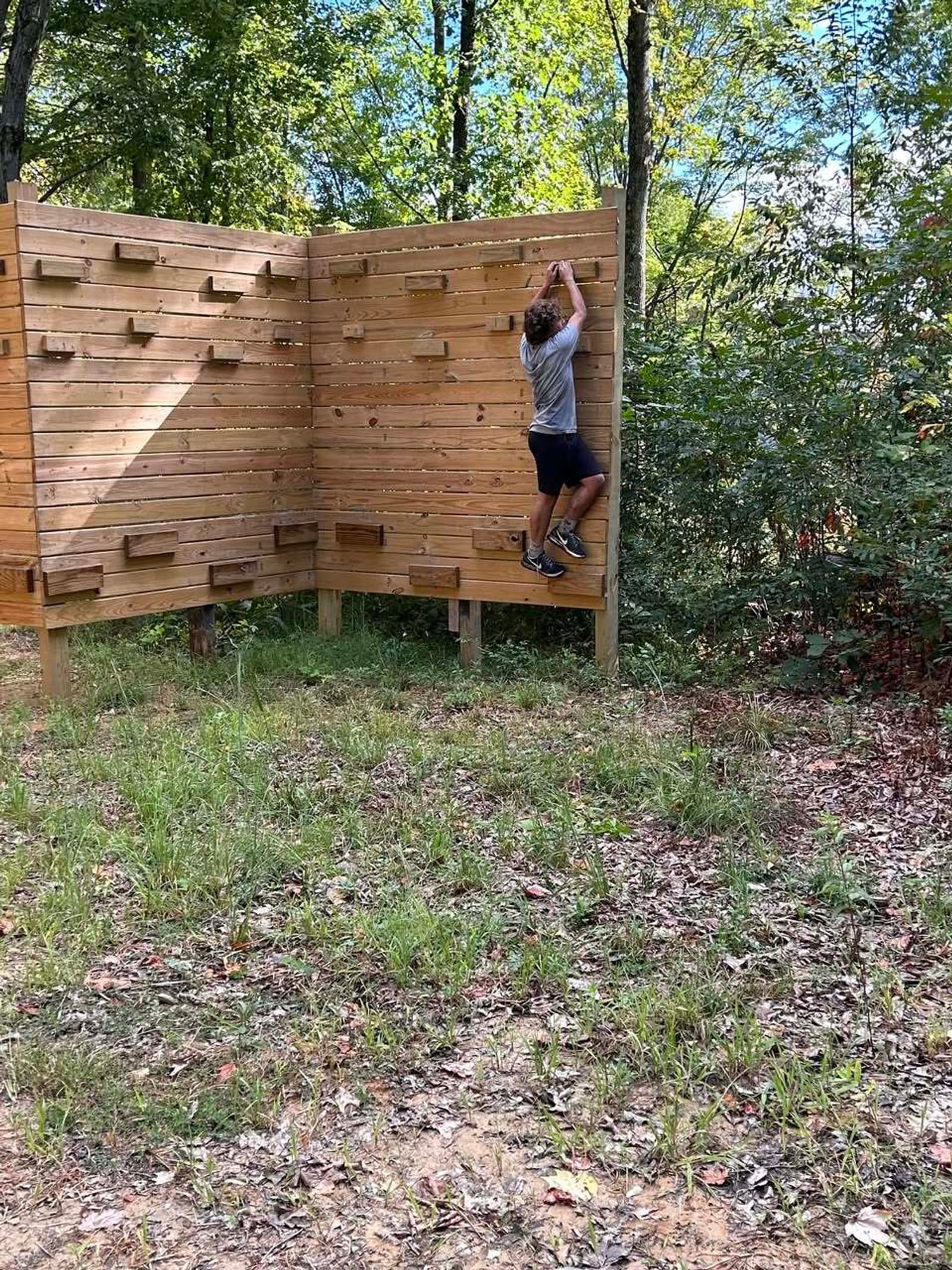 A person climbs a wooden outdoor bouldering wall in a wooded area, gripping the top edge while standing on small handholds. Leafy trees and grass surround the scene.