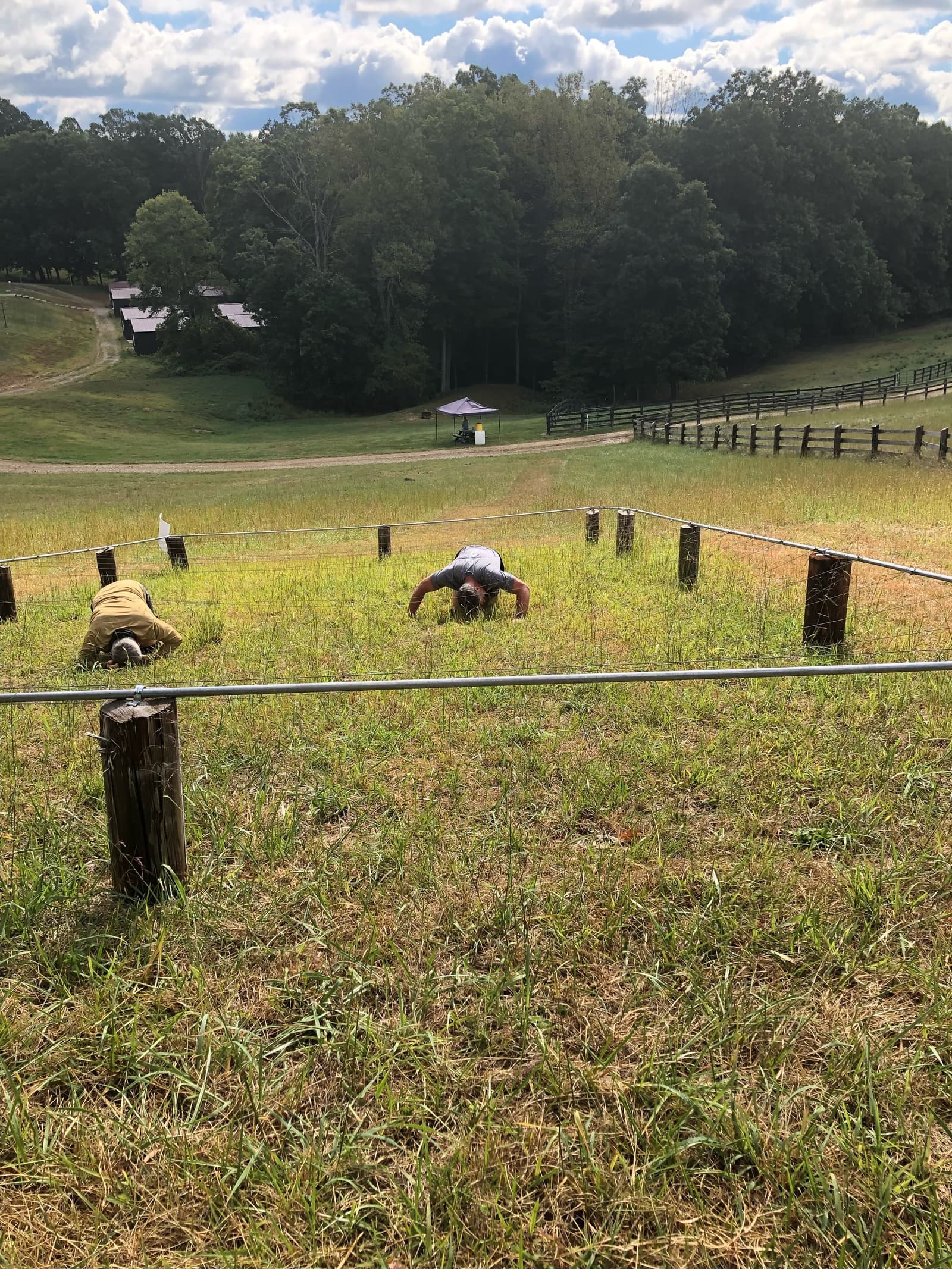 Two people are crawling uphill on grass under a low obstacle made of ropes and wooden posts, with trees and buildings in the background on a partly cloudy day.