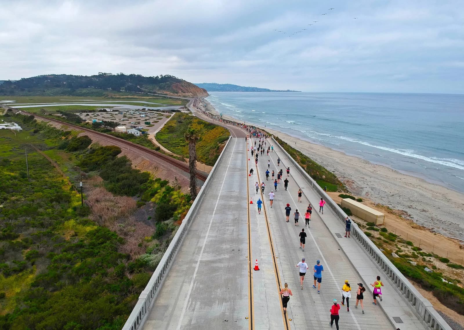 Aerial view of a coastal marathon with participants running along a bridge next to the ocean. Lush greenery is on the left, and the beach is on the right. The sky is cloudy, and hills are visible in the distance.