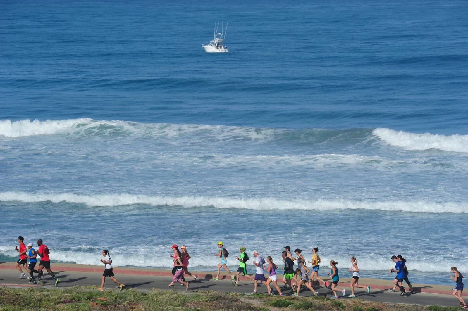 A group of people jogging along a coastal path with the ocean waves in the background. A boat is visible on the sea. The scene suggests a community run or exercise event by the seaside.
