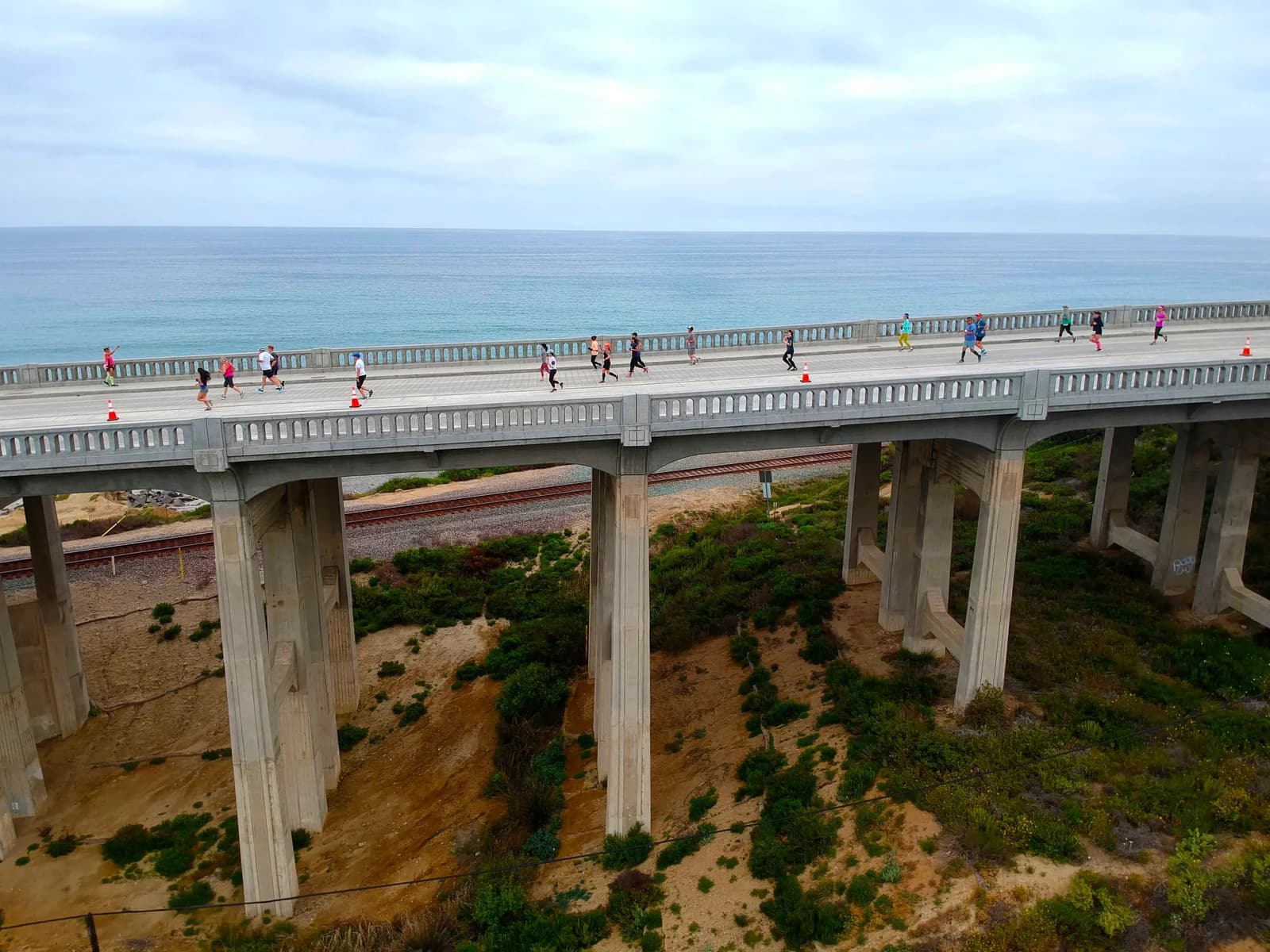 A group of people is running across a long, elevated bridge with railings, overlooking the ocean. The sky is overcast, and there is greenery and a sandy area beneath the bridge.