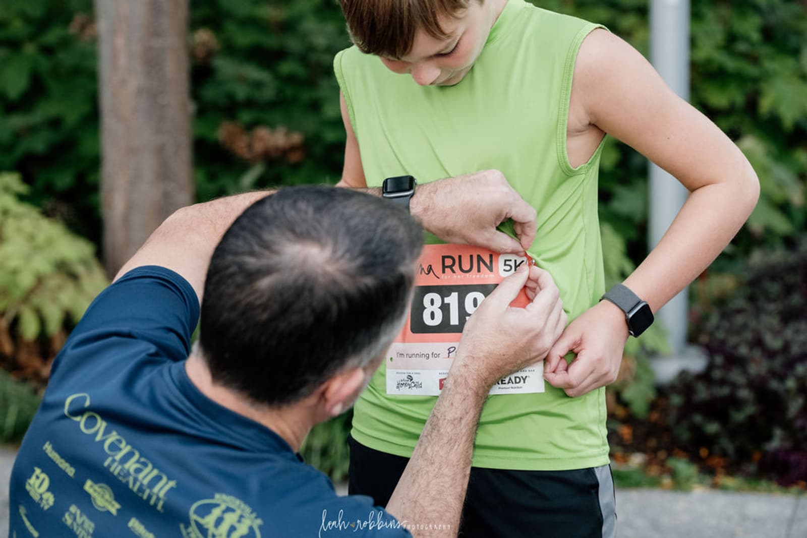 An adult pins a race bib with the number 819 to a child’s green shirt before a 5K run. The child looks down at the bib, and both are outdoors with greenery in the background.