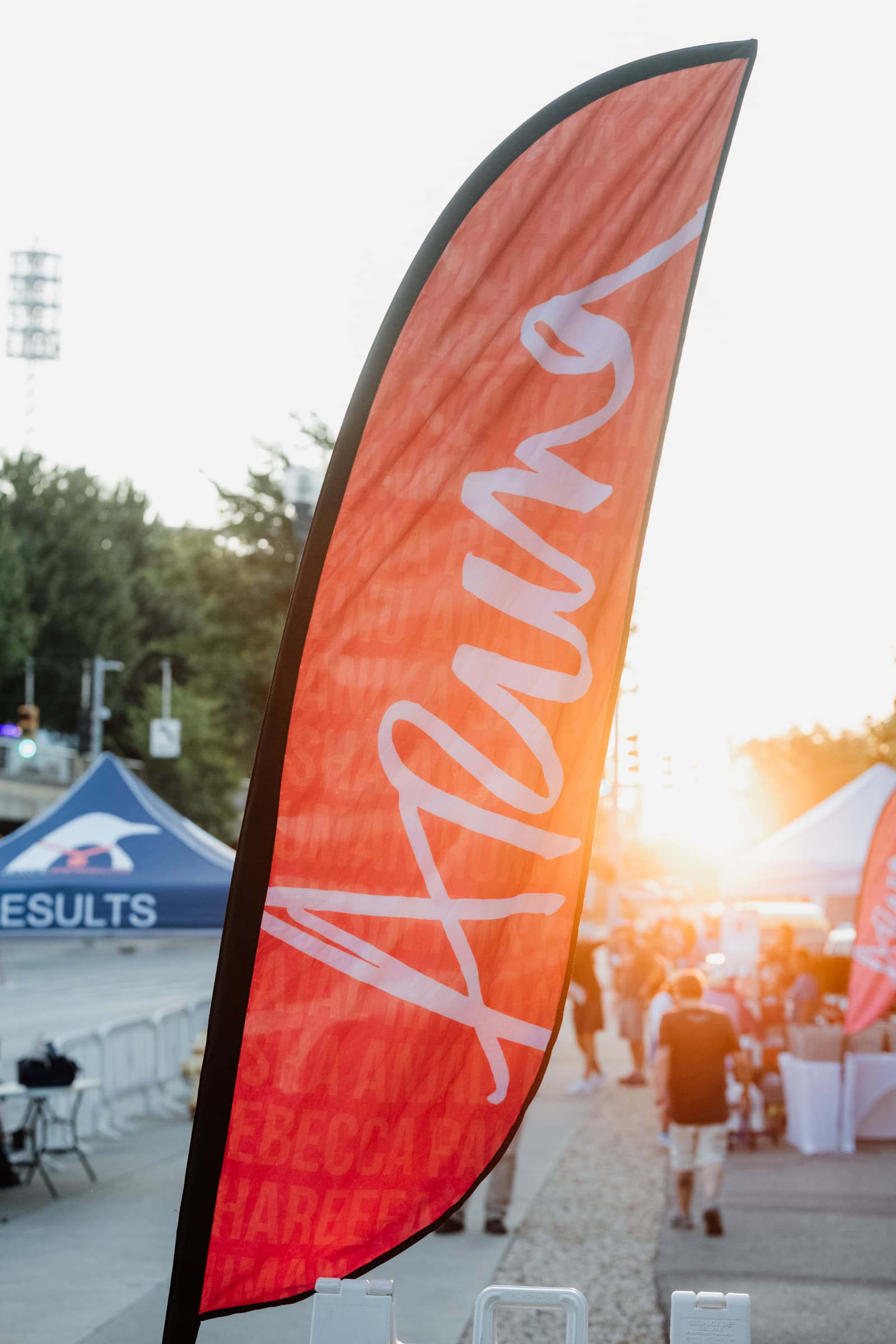 A red vertical flag with the word "RUN" written in large white script letters stands outdoors at an event, with people, tents, and bright sunlight in the background.