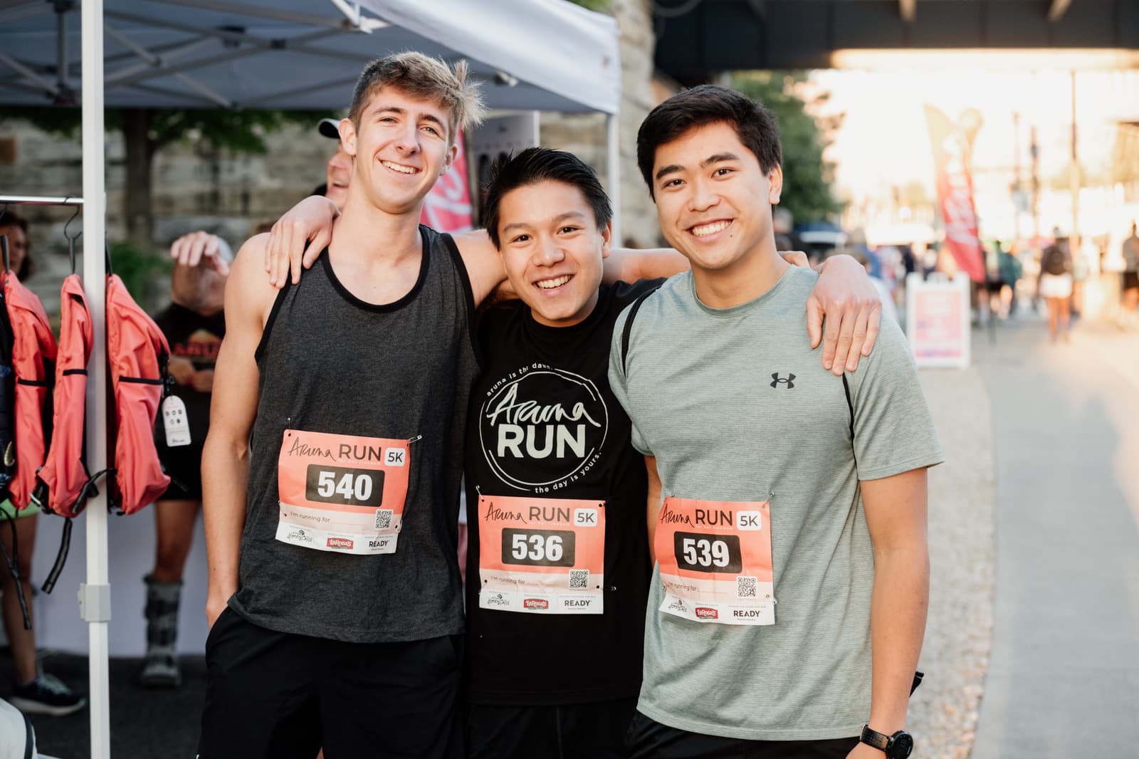 Three young men wearing running gear and race bibs smile with arms around each other at an outdoor running event. A booth with volunteers and safety vests is visible in the background.