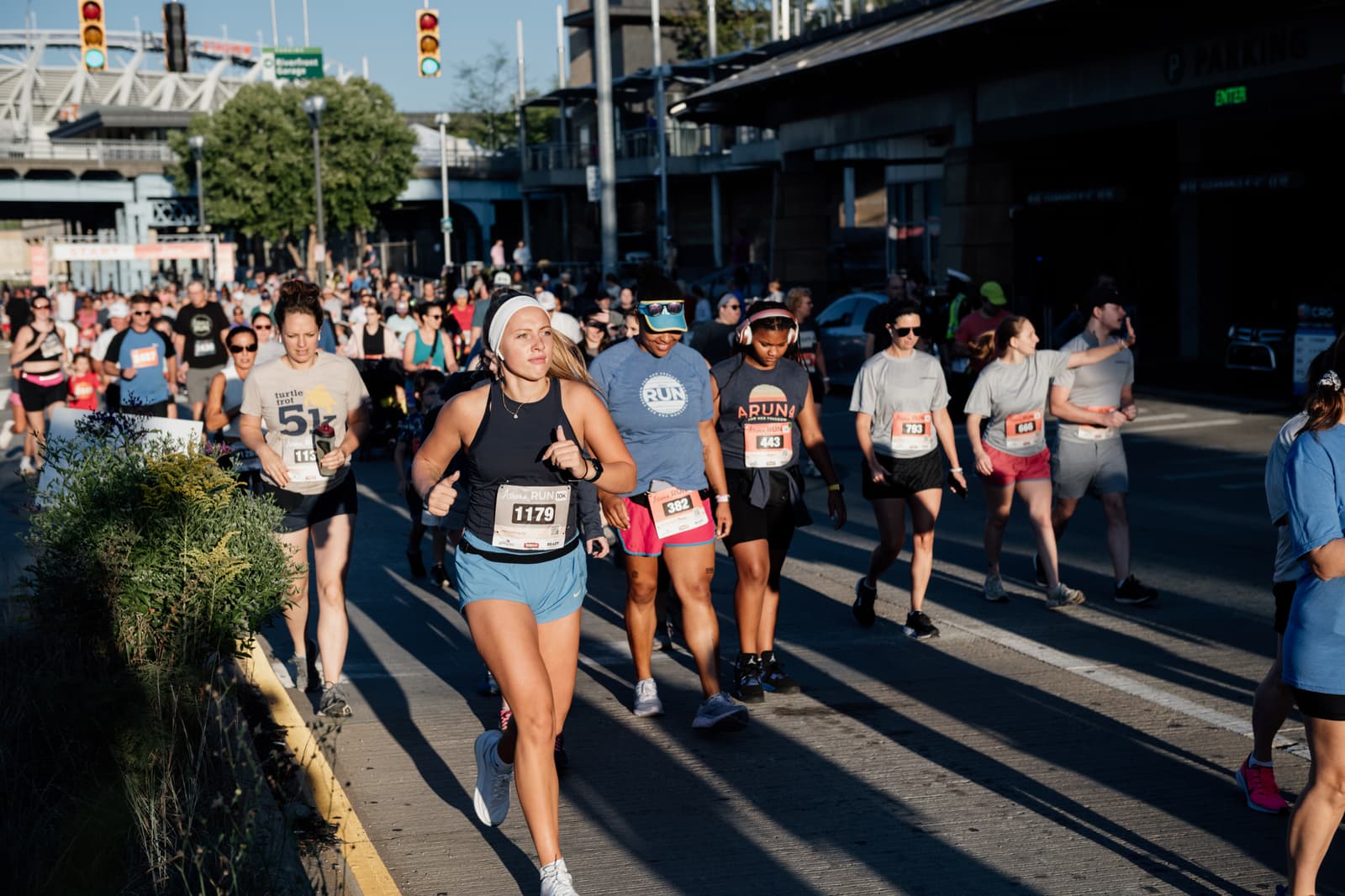 A diverse group of runners, many wearing race bibs, participates in a city 5K event on a sunny day. Some people jog while others walk, and a stadium is visible in the background.