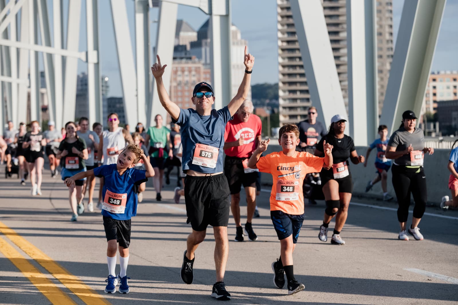 A group of runners crosses a bridge during a race. A man in sunglasses raises his arms in celebration, running alongside two smiling boys. Other participants follow behind, city buildings visible in the background.