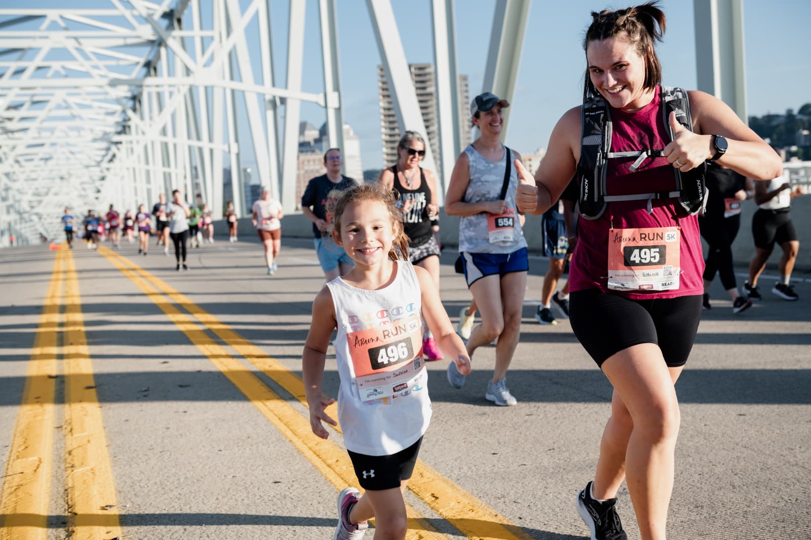 A smiling young girl and an enthusiastic woman run in a race on a bridge with other participants behind them; both wear race bibs and athletic clothing in bright sunlight.