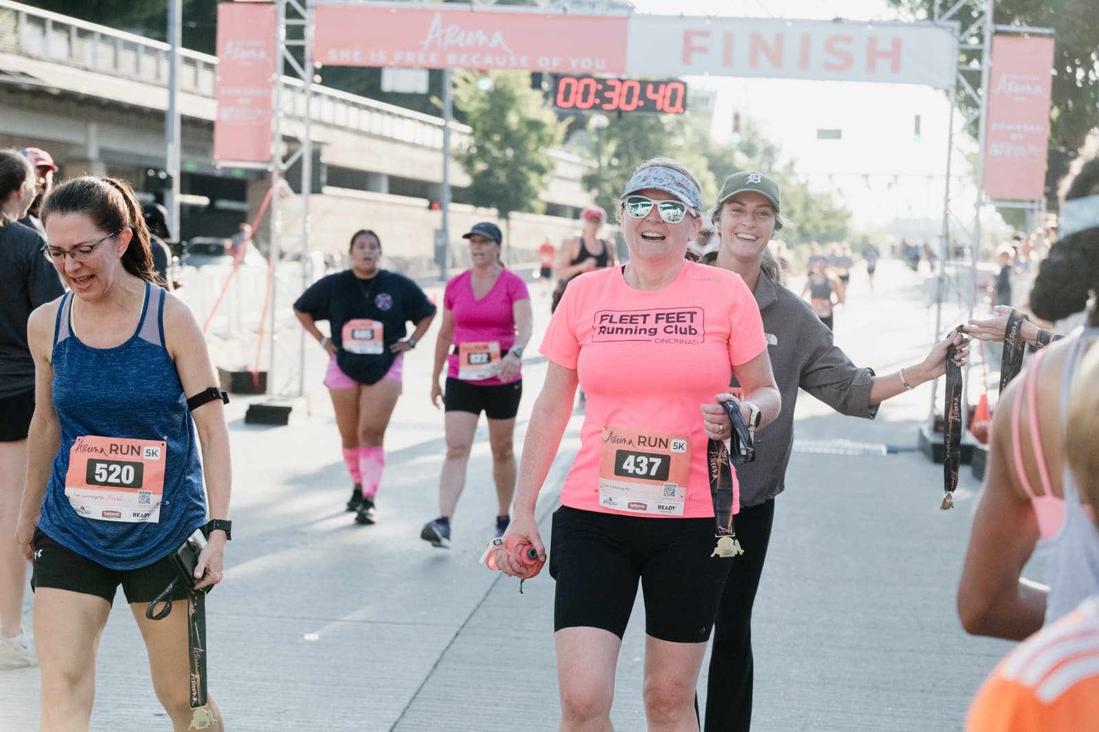 Runners smiling and walking past the finish line at a race event. The digital timer shows 30:40. One woman in a bright pink "Fleet Feet Running Club" shirt stands out, holding a medal and looking happy. Other runners are in the background.