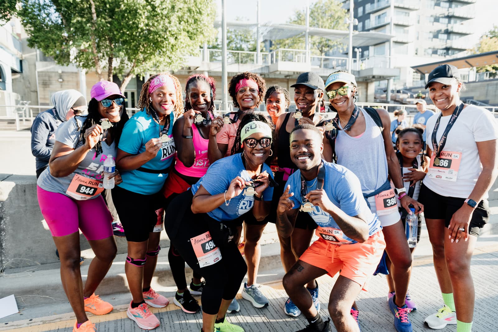 A group of smiling runners pose together outdoors after a race, proudly displaying their medals. Most are wearing athletic gear and numbered bibs. The mood is joyful and celebratory on a sunny day.