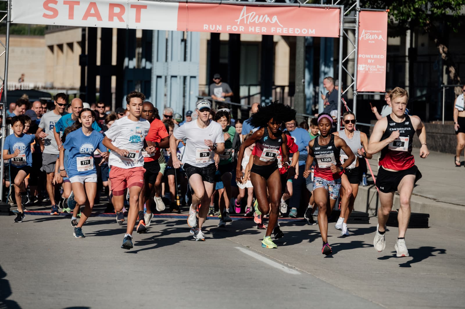 A diverse group of runners in athletic gear start a race on a city street, passing under a banner that reads "RUN FOR HER FREEDOM" with the word "START" above them. Spectators and buildings are visible in the background.