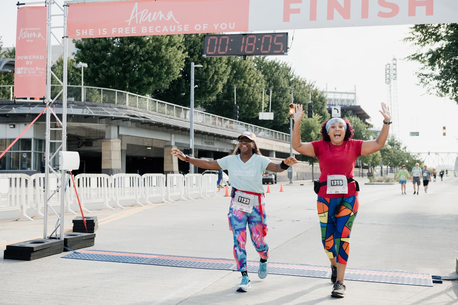 Two women with race bibs joyfully cross a finish line outdoors, raising their arms in celebration under a sign that reads βSHE IS FREE BECAUSE OF YOU.β A race timer above them shows 18:05.