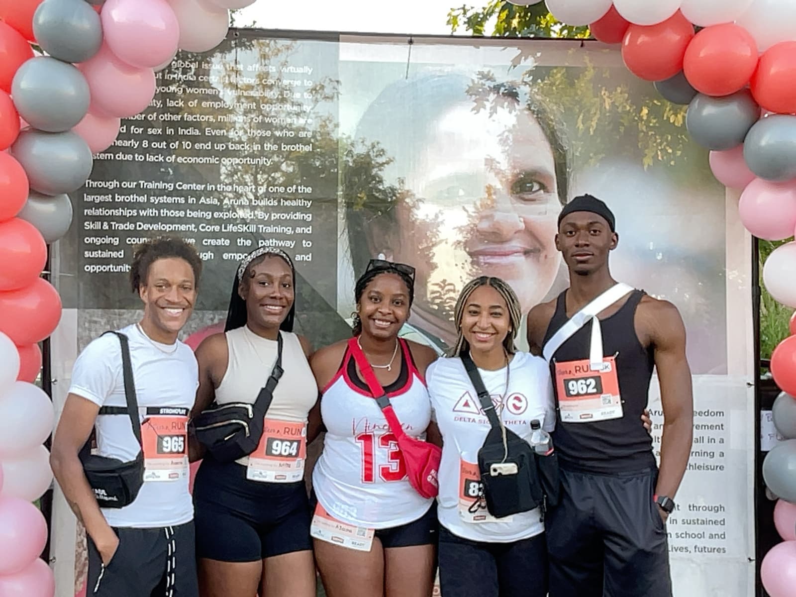 Five smiling young adults stand together under a red, white, and gray balloon arch, wearing running gear and race bibs. Behind them is a large banner with text and an image of a person’s face.