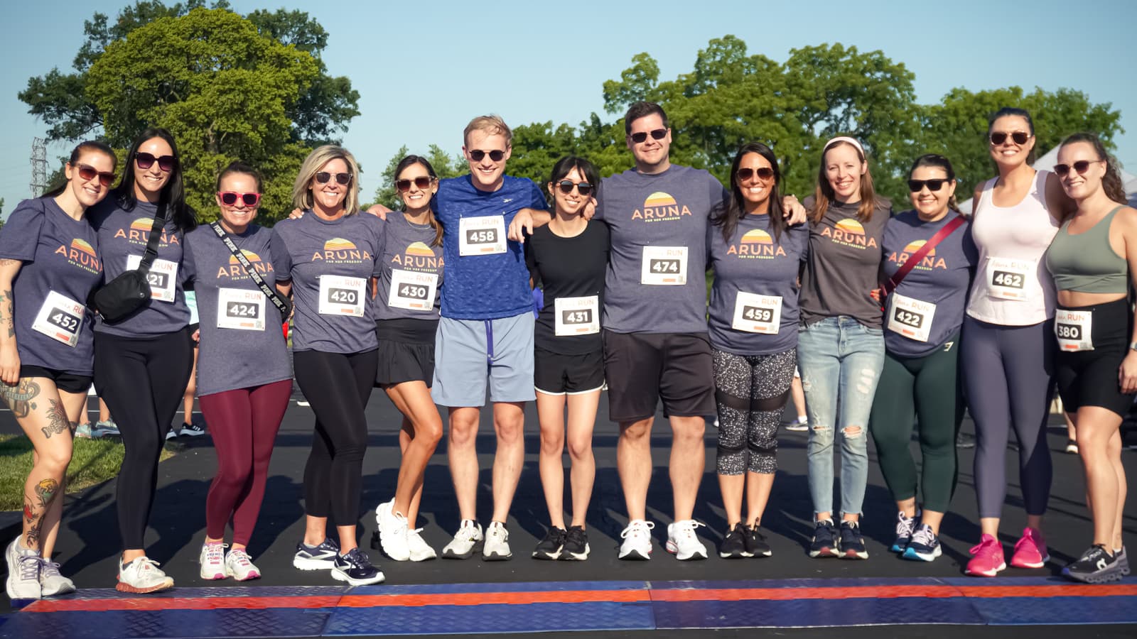 A group of runners smile and pose together outdoors before a race, wearing numbered bibs and athletic gear. Some wear matching “Aruna” T-shirts, and the weather is sunny with green trees in the background.