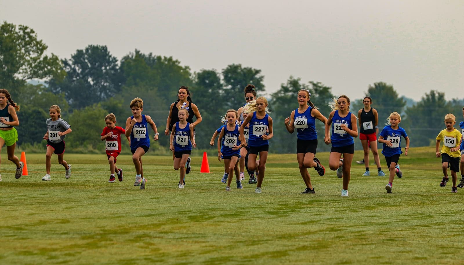 A group of children and teens wearing athletic clothes and race bibs run on a grassy field during a cross-country race, with orange cones and trees visible in the background.