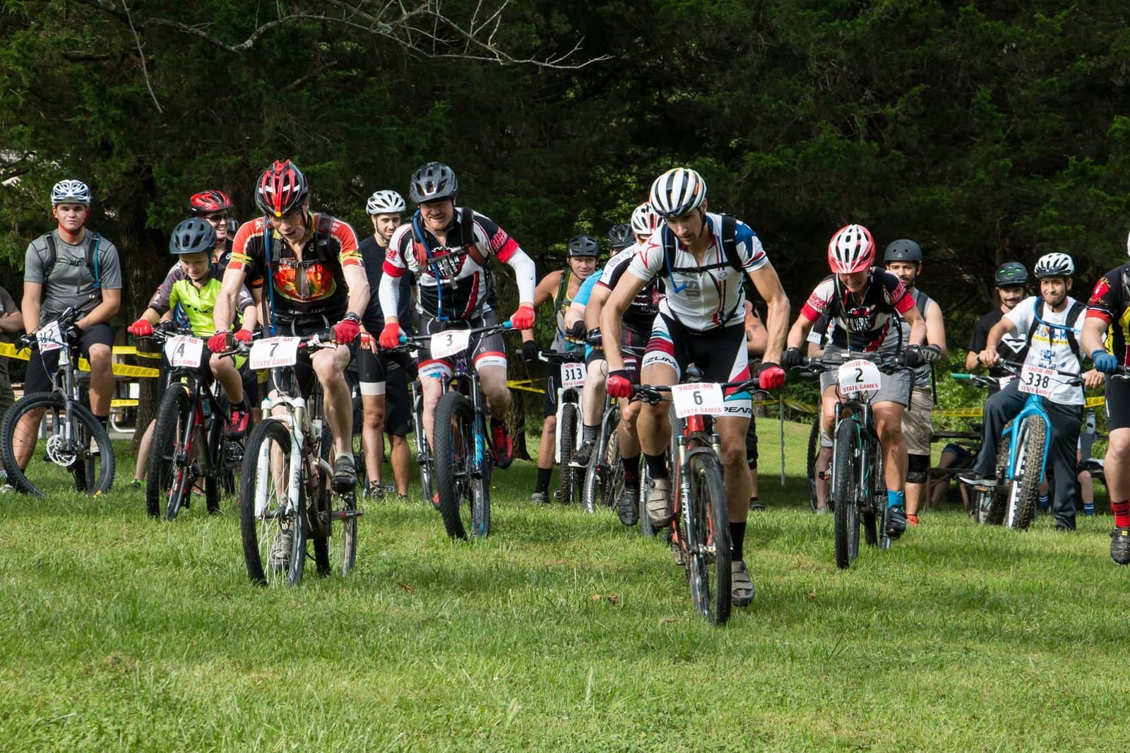 A group of cyclists wearing helmets and numbered jerseys ride mountain bikes on grass, starting a race. Trees and other racers are visible in the background.