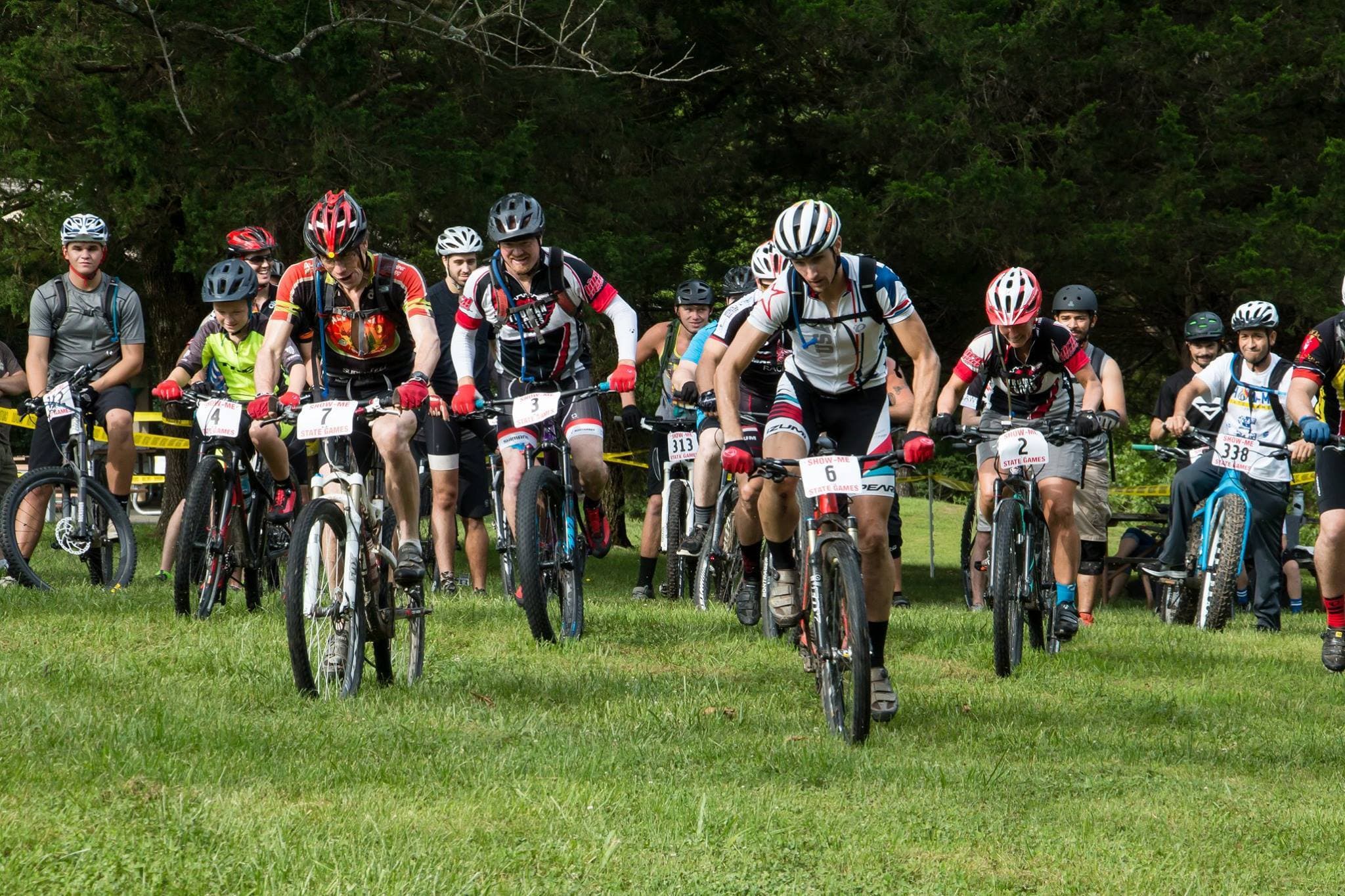 A group of cyclists wearing helmets and numbered jerseys ride mountain bikes on grass, starting a race. Trees and other racers are visible in the background.