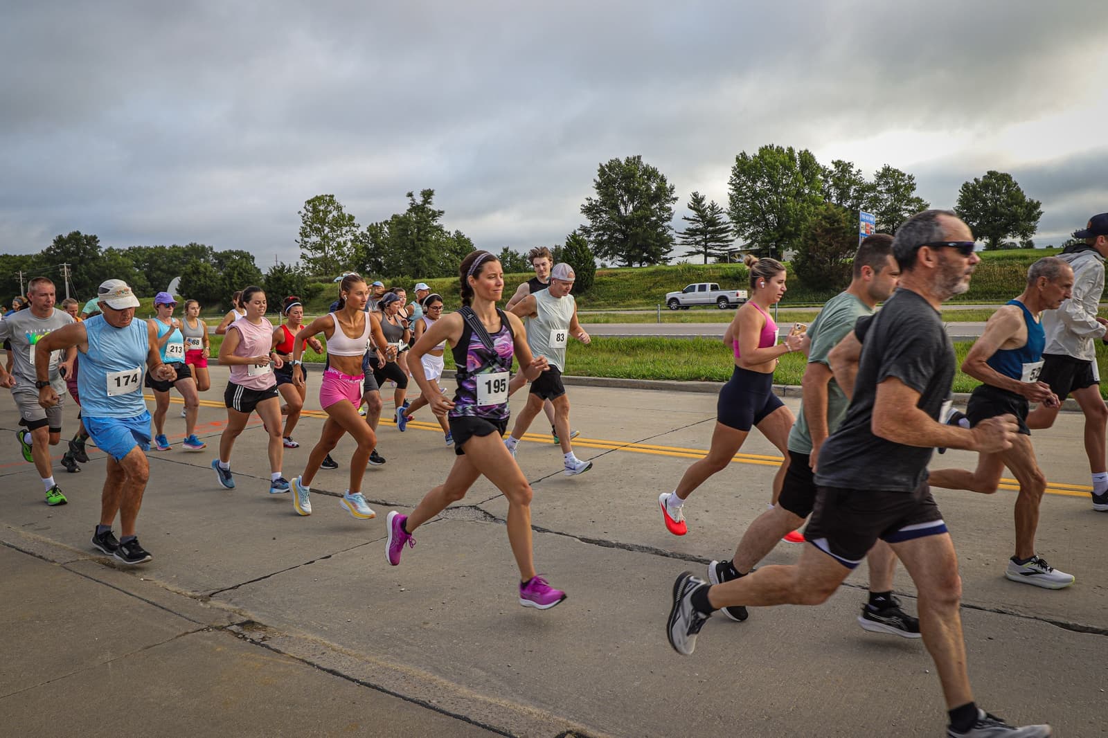 A group of runners of various ages and genders participates in a road race on a cloudy day, running along a paved street surrounded by trees and grass. Some runners wear bib numbers and athletic gear.