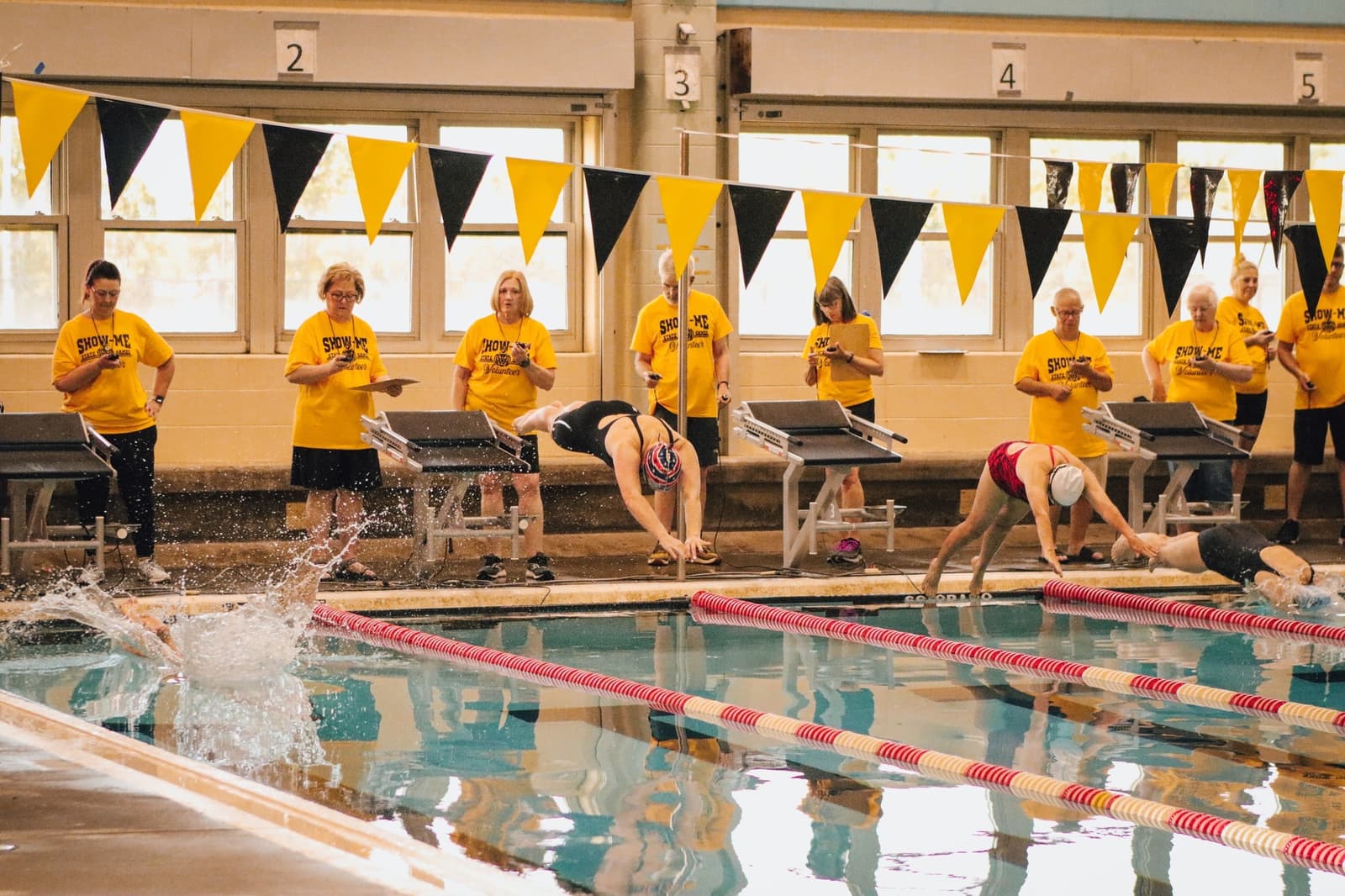 Three swimmers dive into an indoor pool at the start of a race, while a group of people in yellow shirts labeled “SWIM” watch from behind the lanes. Black and yellow flags hang above the pool.
