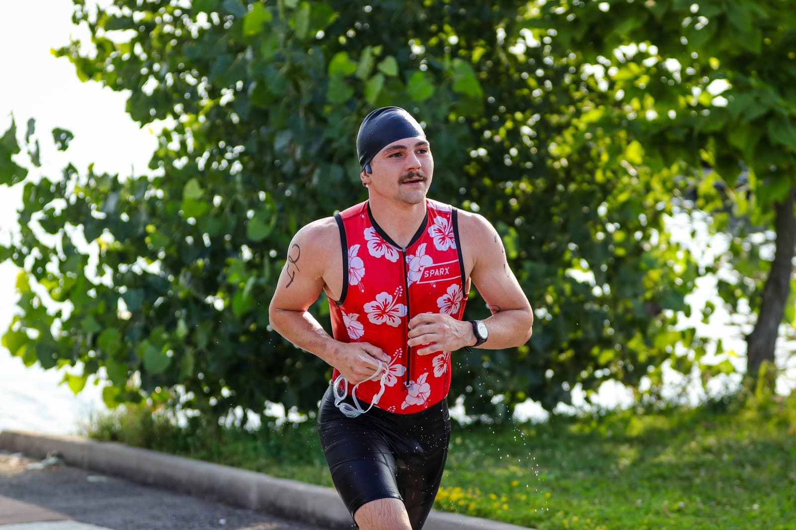 A man wearing a red, floral-patterned triathlon suit and a black swim cap runs outdoors on a sunny day, holding goggles in one hand. Lush green trees and a lake are visible in the background.