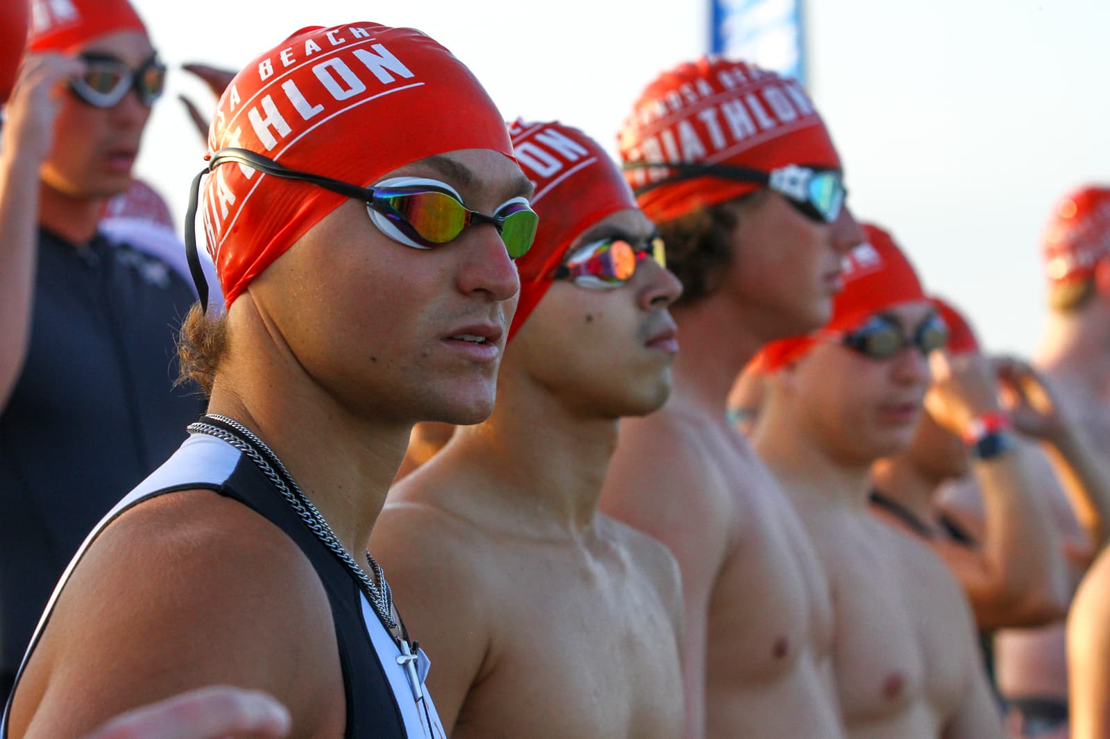 A group of athletes wearing red swim caps and goggles stand at the starting line of a triathlon, looking focused and ready to begin the race.