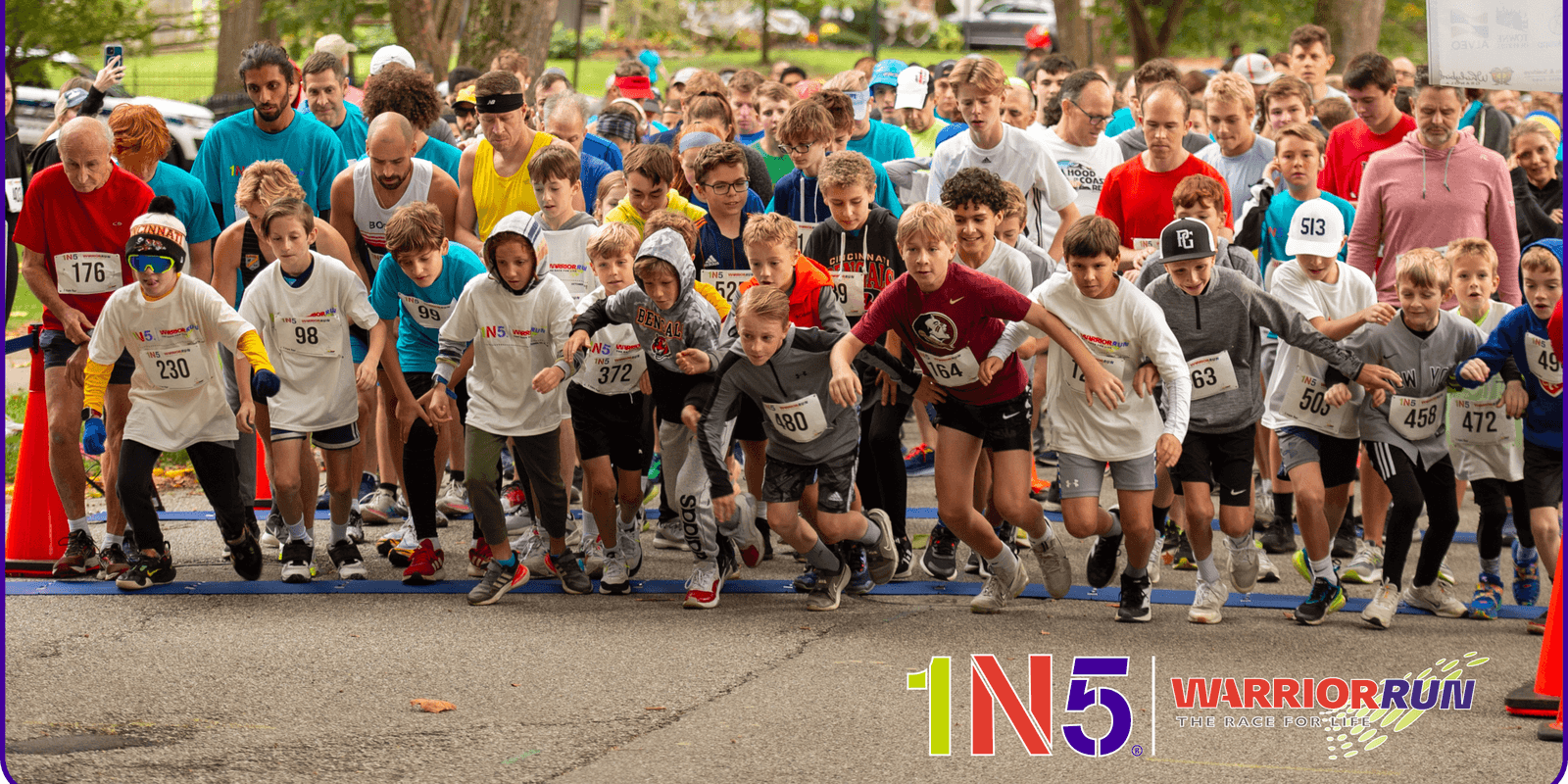 A large, diverse group of runners of various ages begin a race outdoors on a paved path, with “1N5 Warrior Run” logos in colorful letters visible at the bottom of the image.