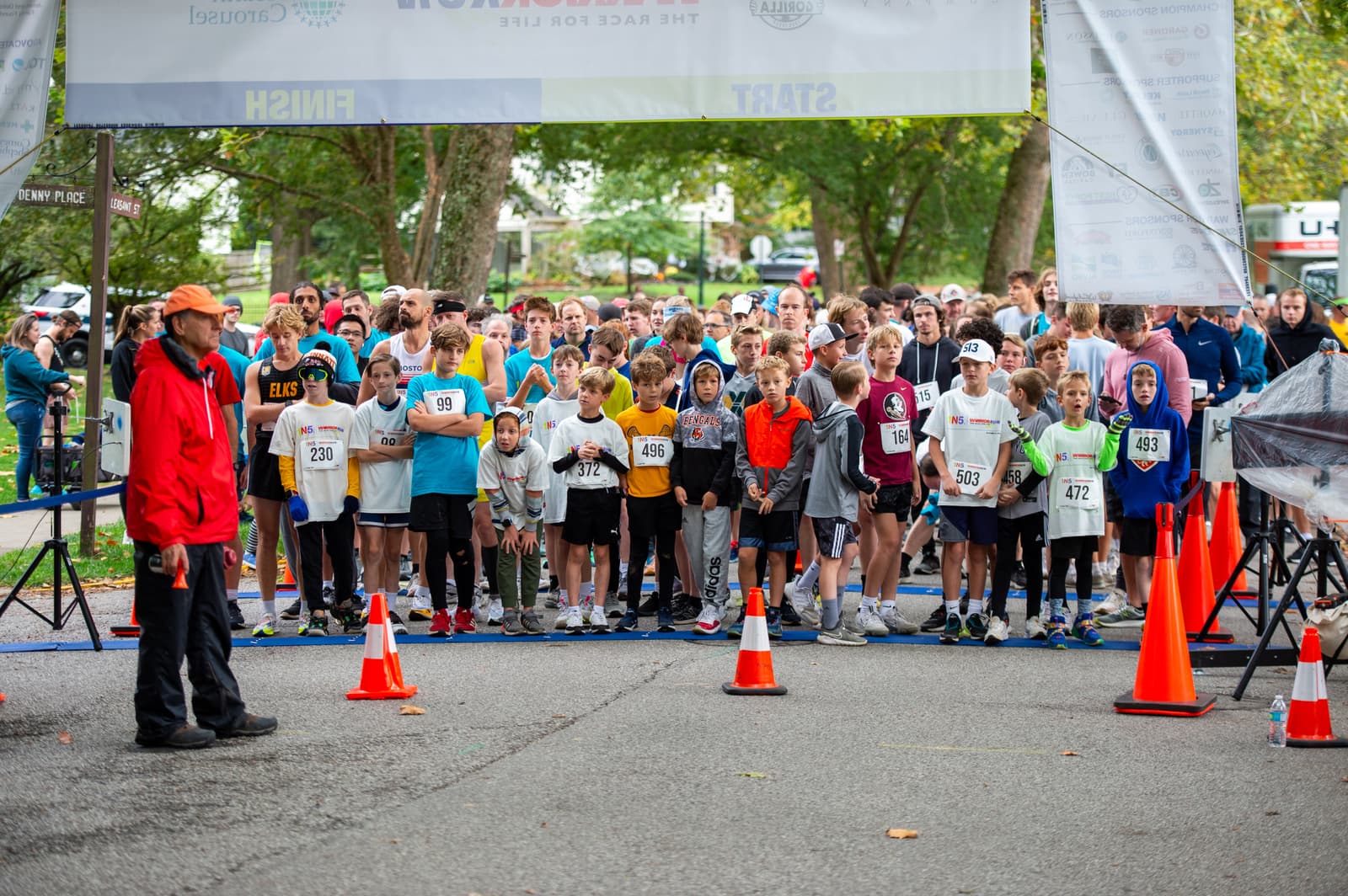 A large group of people, including children and adults, stand at the starting line of a race, ready to run. A person in a red jacket stands in front, and orange cones line the path. Trees and banners are visible in the background.
