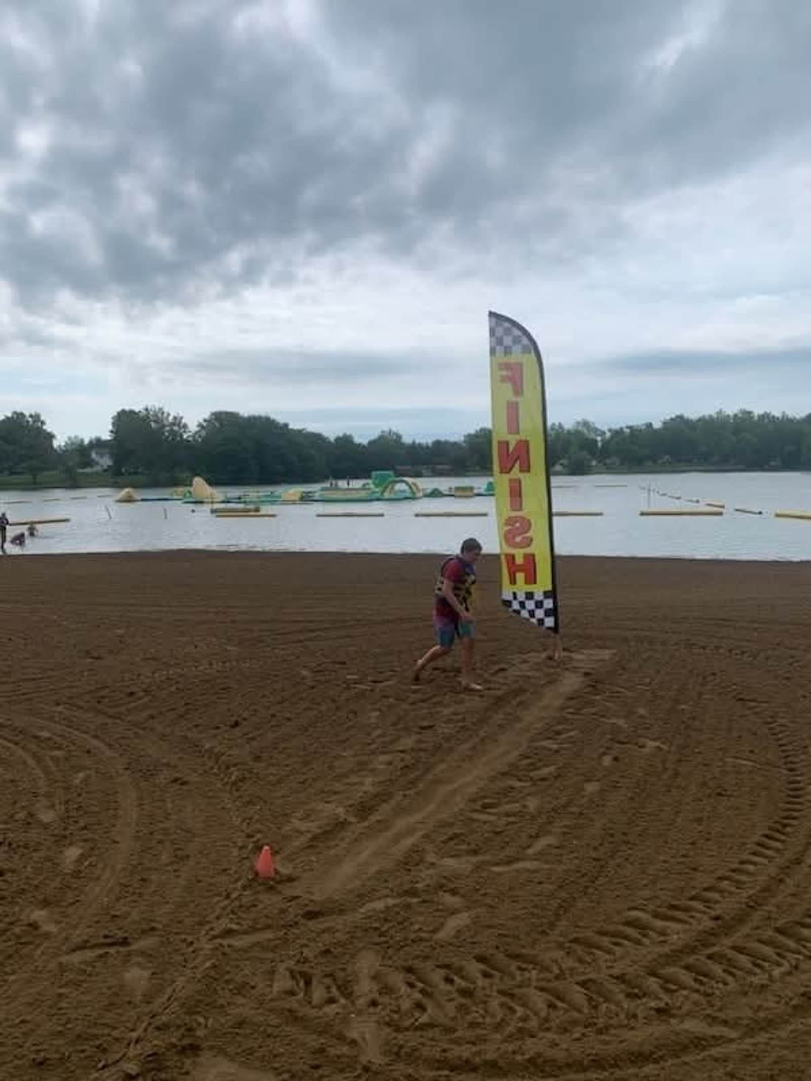 A child wearing swim gear walks past a tall "FINISH" flag on a sandy beach near a lake, with inflatables visible in the water and a cloudy sky overhead.