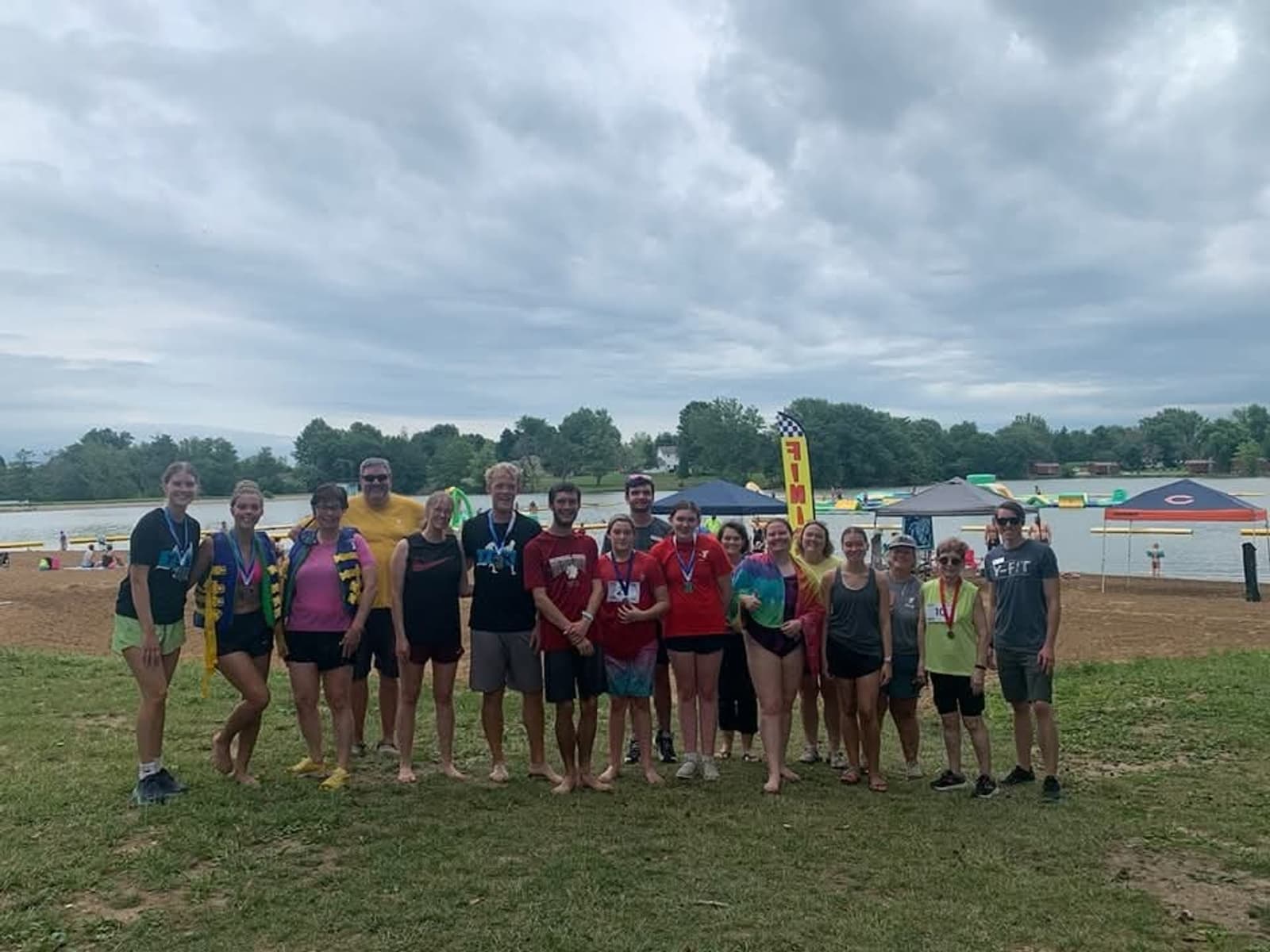 A group of people stand barefoot on grass near a lake, smiling at the camera. Colorful banners, tents, and inflatables are visible in the background under a cloudy sky.