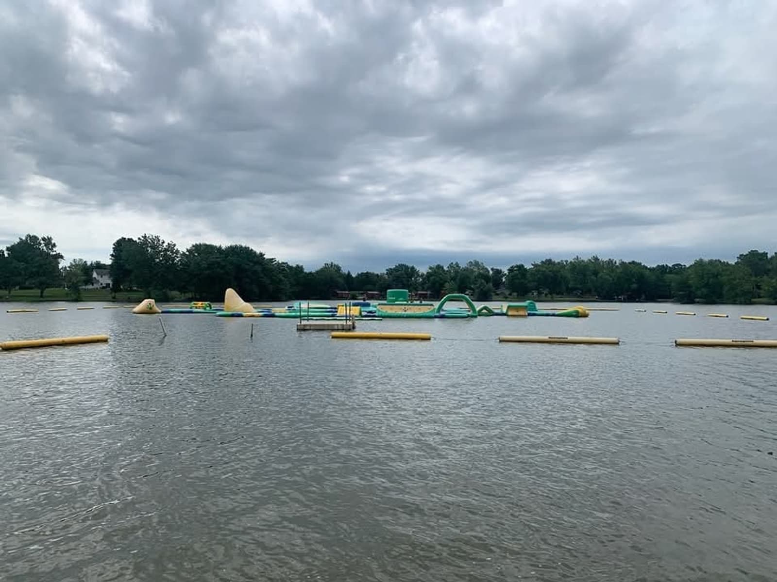 A lake with floating yellow barriers and an inflatable water obstacle course, surrounded by trees under a cloudy sky.