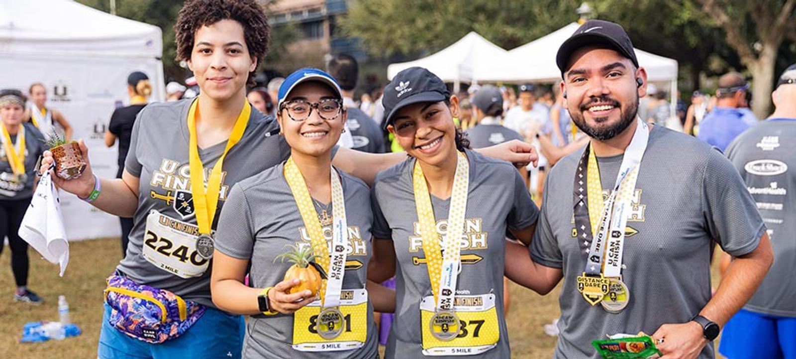 Four smiling runners wearing medals and race bibs pose together at a finish line event, holding snacks and drinks. White tents and other participants are visible in the background.