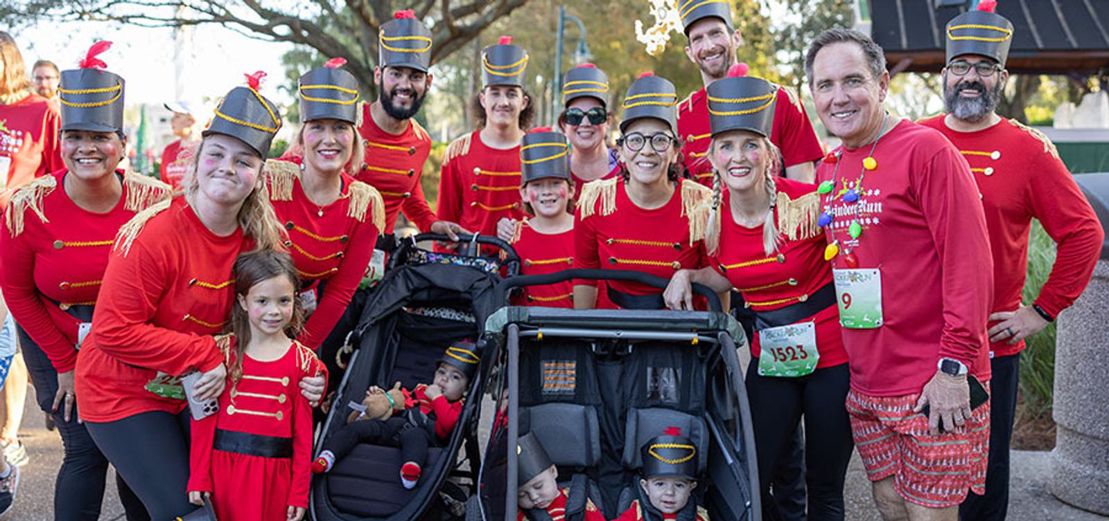 A group of adults and children, many in matching red toy soldier costumes with hats, smile at the camera outdoors. Two toddlers sit in strollers dressed in similar costumes. Trees and sunlight are in the background.