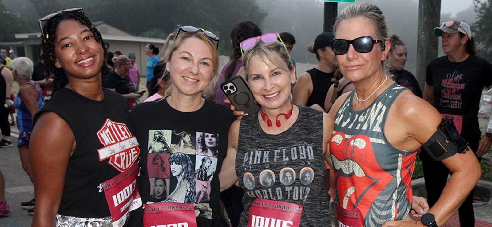Four women stand close together outdoors at a race event, smiling at the camera. They wear race bibs and rock band shirts. Other participants and trees are visible in the background.