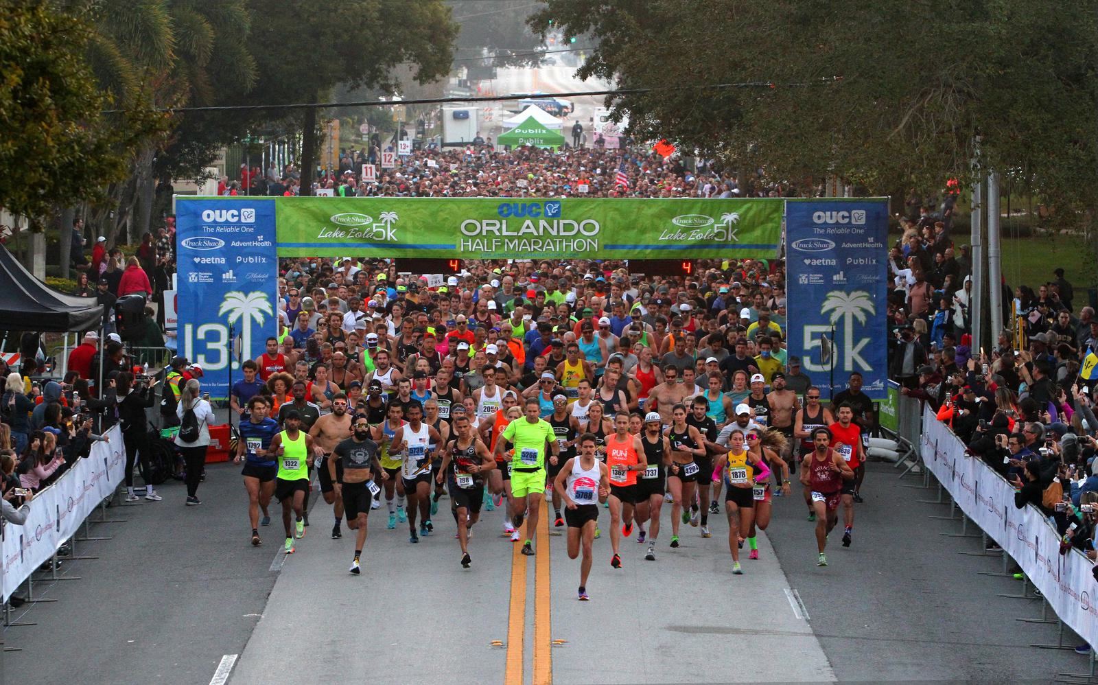 A large crowd of runners starts the OUC Orlando Half Marathon and 5K race, passing under a green banner, with spectators lining both sides of the street and trees in the background.