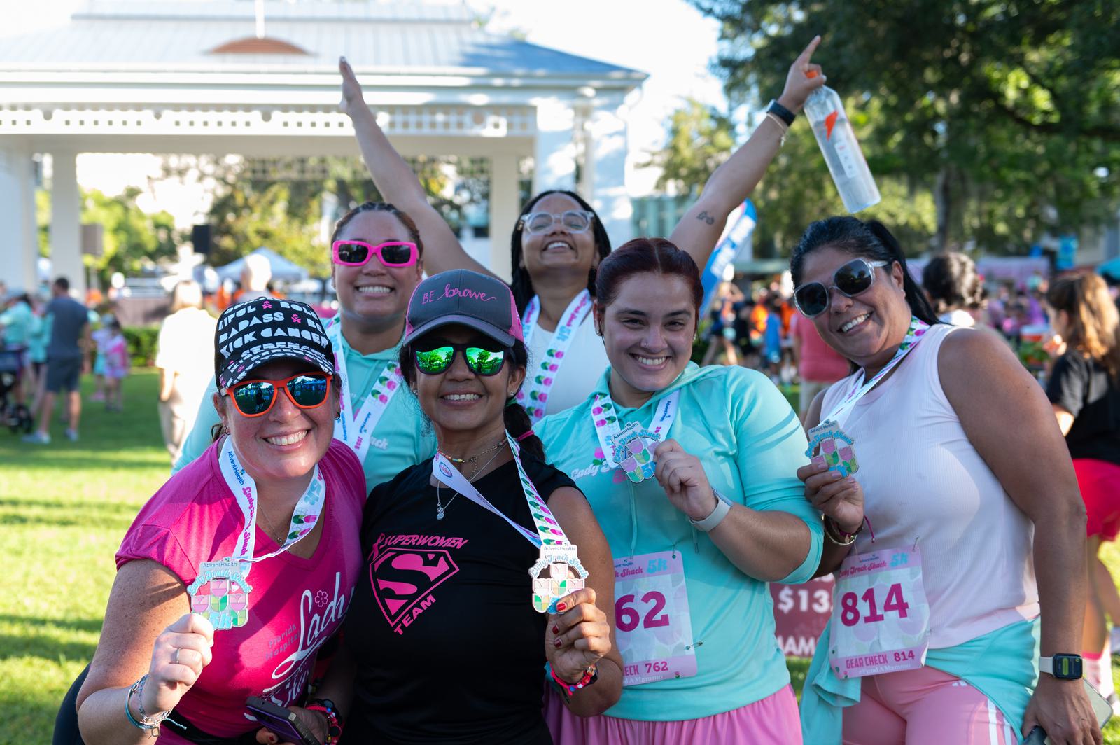A group of six smiling women wearing athletic gear and race bibs pose together in a park, proudly showing off their medals after a race event. The atmosphere is joyful and celebratory.