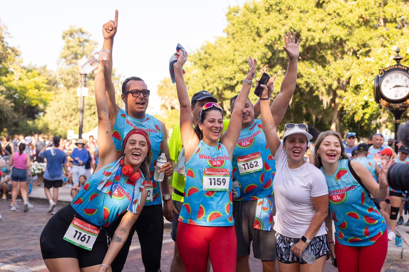 A group of happy runners in matching watermelon-themed outfits celebrate with raised arms after finishing a race, smiling at the camera in a lively outdoor setting with trees and other participants in the background.
