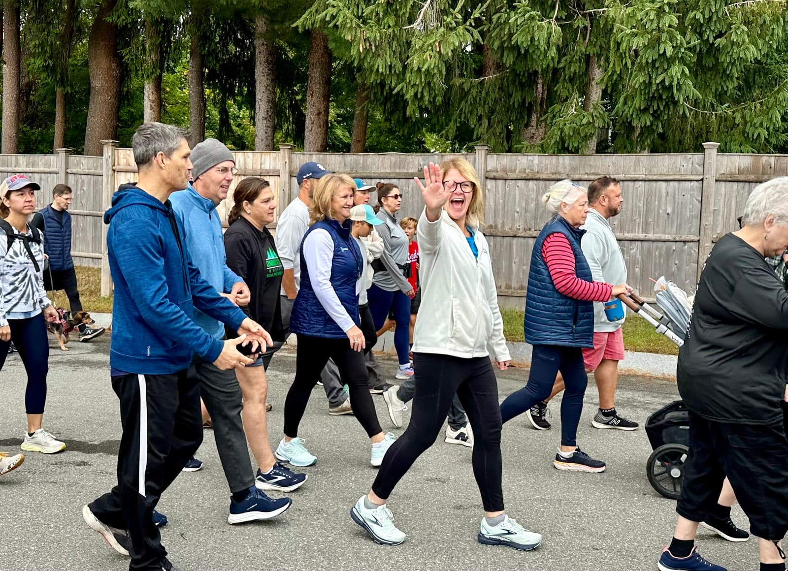 A group of people, dressed in casual sportswear, walk together outdoors near a wooden fence. One woman in the center waves and smiles at the camera; a stroller and trees are visible in the background.