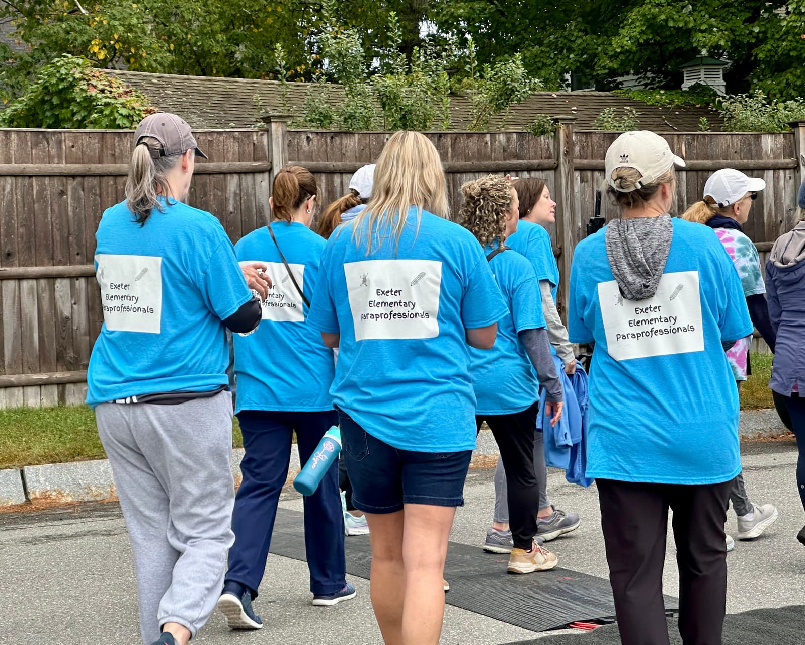 A group of people wearing matching blue "Exeter Elementary Paraprofessionals" shirts and baseball caps walk together outdoors near a wooden fence.