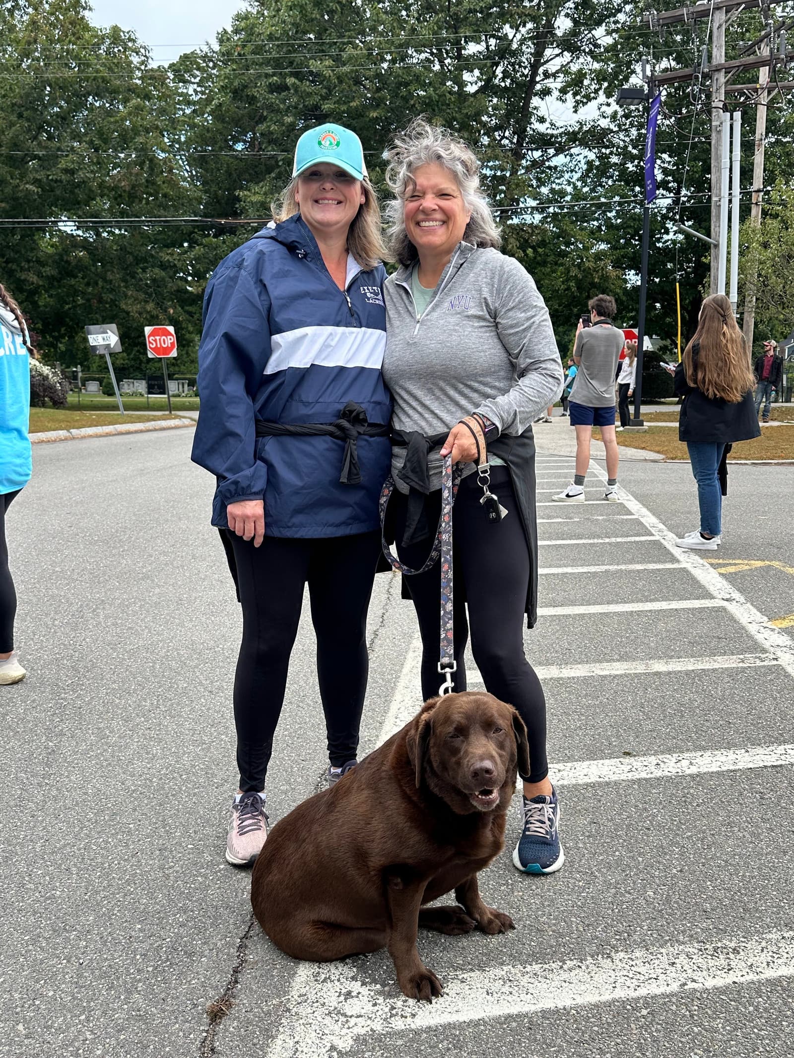 Two women stand smiling in a parking lot, one holding the leash of a large brown dog sitting in front of them. Several people are visible in the background near trees and a street.