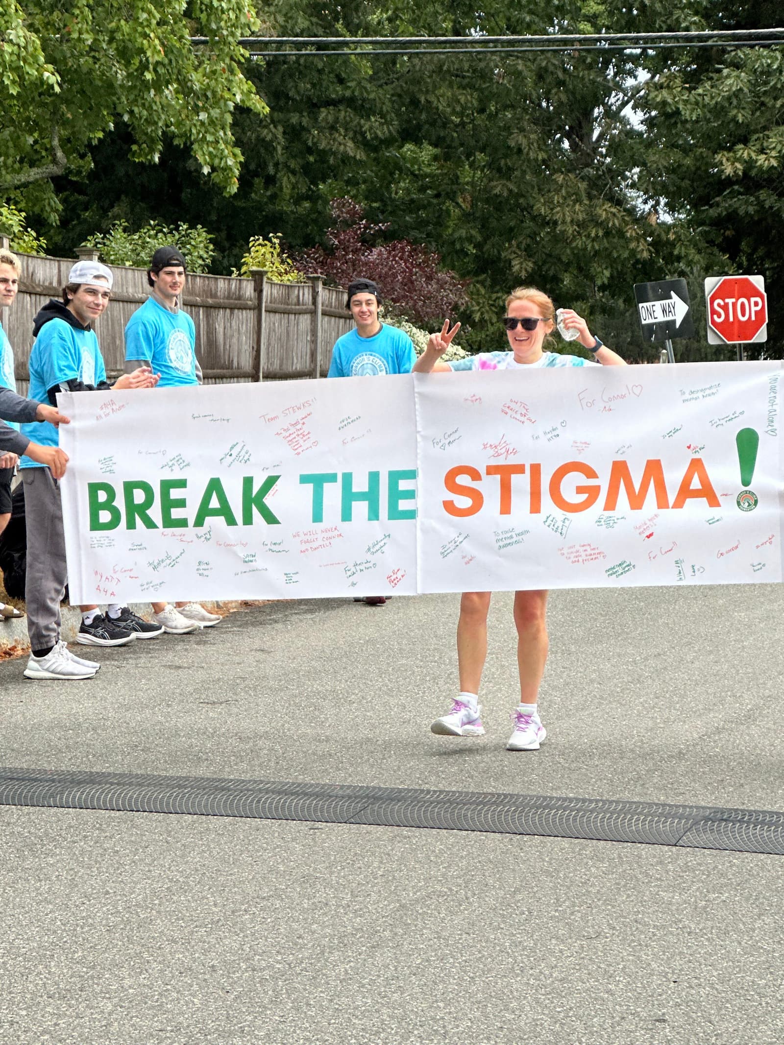 A group of people in blue shirts stand on a street holding a large banner that reads "BREAK THE STIGMA!" A smiling woman in sunglasses stands in front of the banner making a peace sign. Trees and a stop sign are in the background.